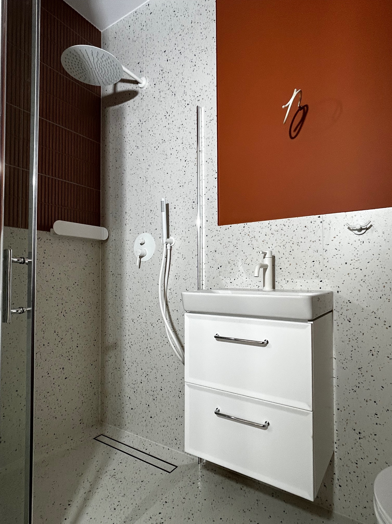 Nowoczesna łazienka z kabiną prysznicową typu walk-in, umywalką z szafką w kolorze białym i ścianami wykończonymi płytkami lastryko. Akcent stanowi pomarańczowa ściana.