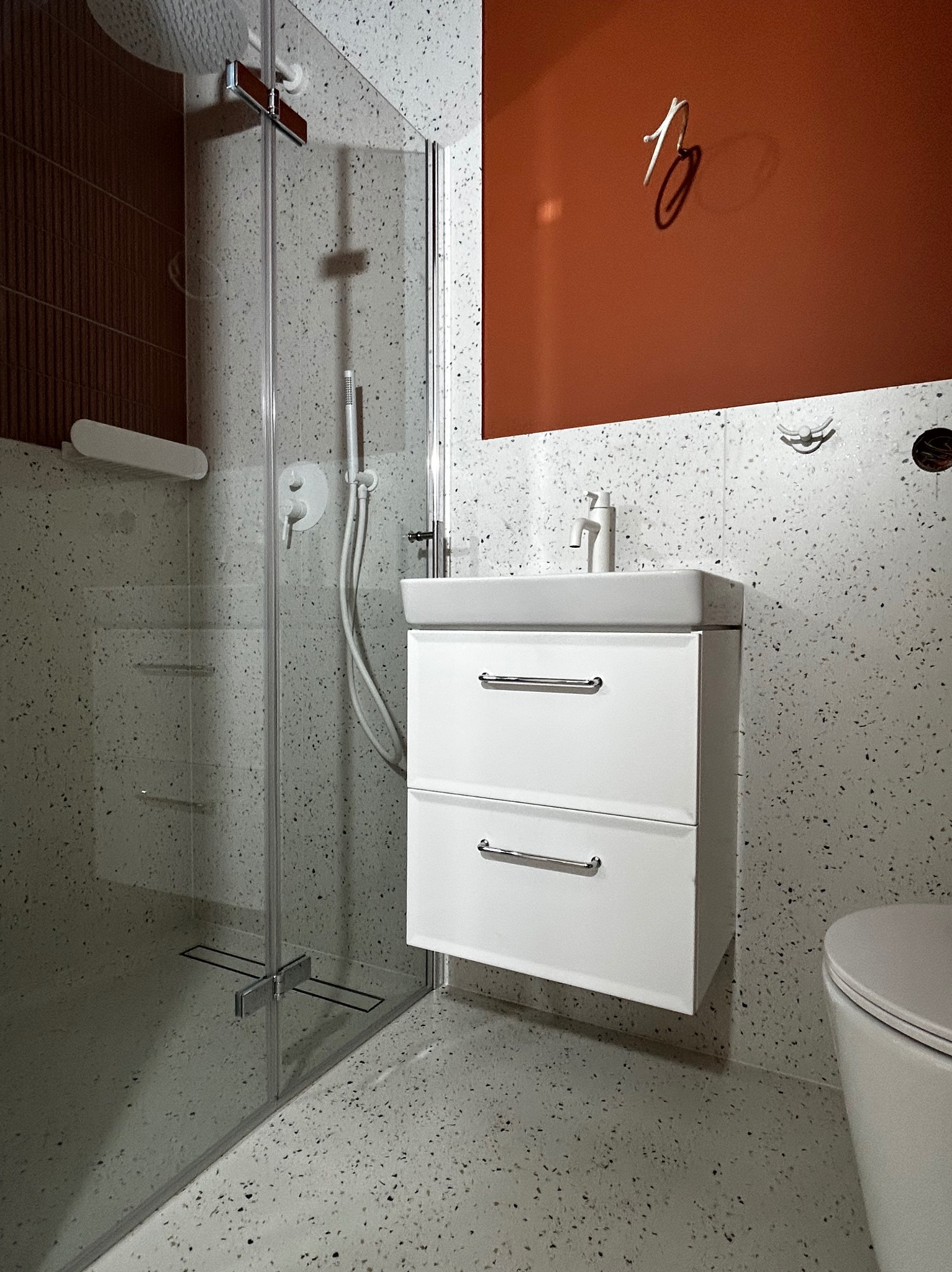 Nowoczesna łazienka z kabiną prysznicową typu walk-in, białą szafką pod umywalką i ścianami wykończonymi w stylu lastryko. Akcent stanowi pomarańczowa ściana.