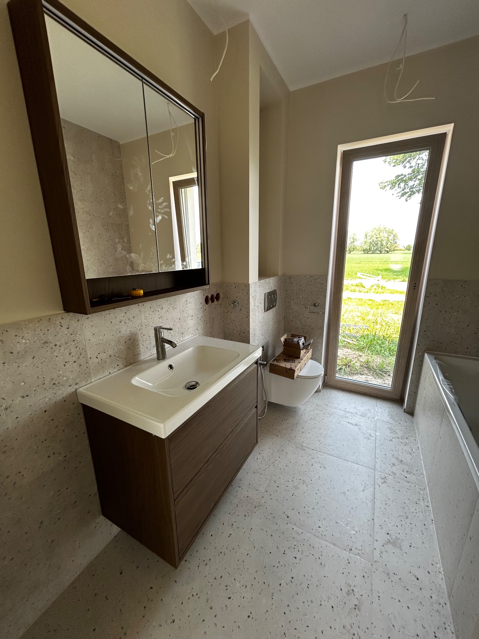 Nowoczesna łazienka z umywalką w szafce z ciemnego drewna, lustrem z szafką, toaletą i oknem z widokiem na zieleń. Płytki w jasnym kolorze z ciemnymi drobinkami.