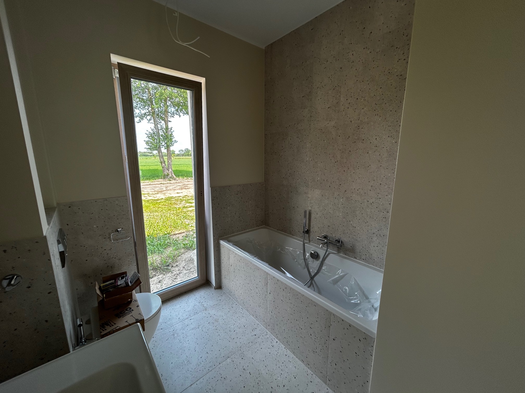 Jasna łazienka z oknem na zielony krajobraz. Wanna obudowana płytkami w kolorze ścian. Umywalka i toaleta częściowo widoczne. Minimalistyczny design.