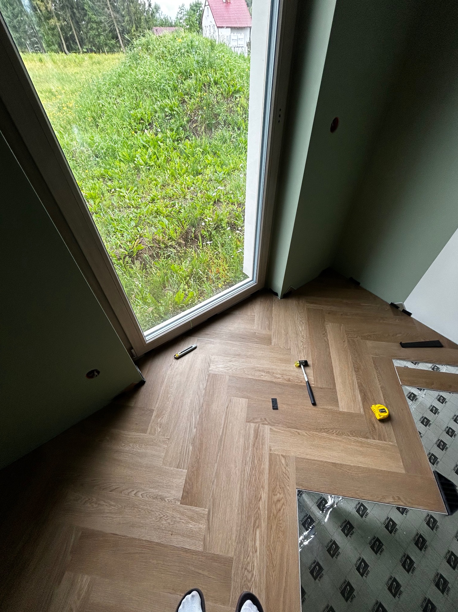 Układanie parkietu w jodełkę w pokoju z oknem na ogród. Widoczne narzędzia: młotek, miarka, nóż. Częściowo ułożona podłoga, panele z folią ochronną.