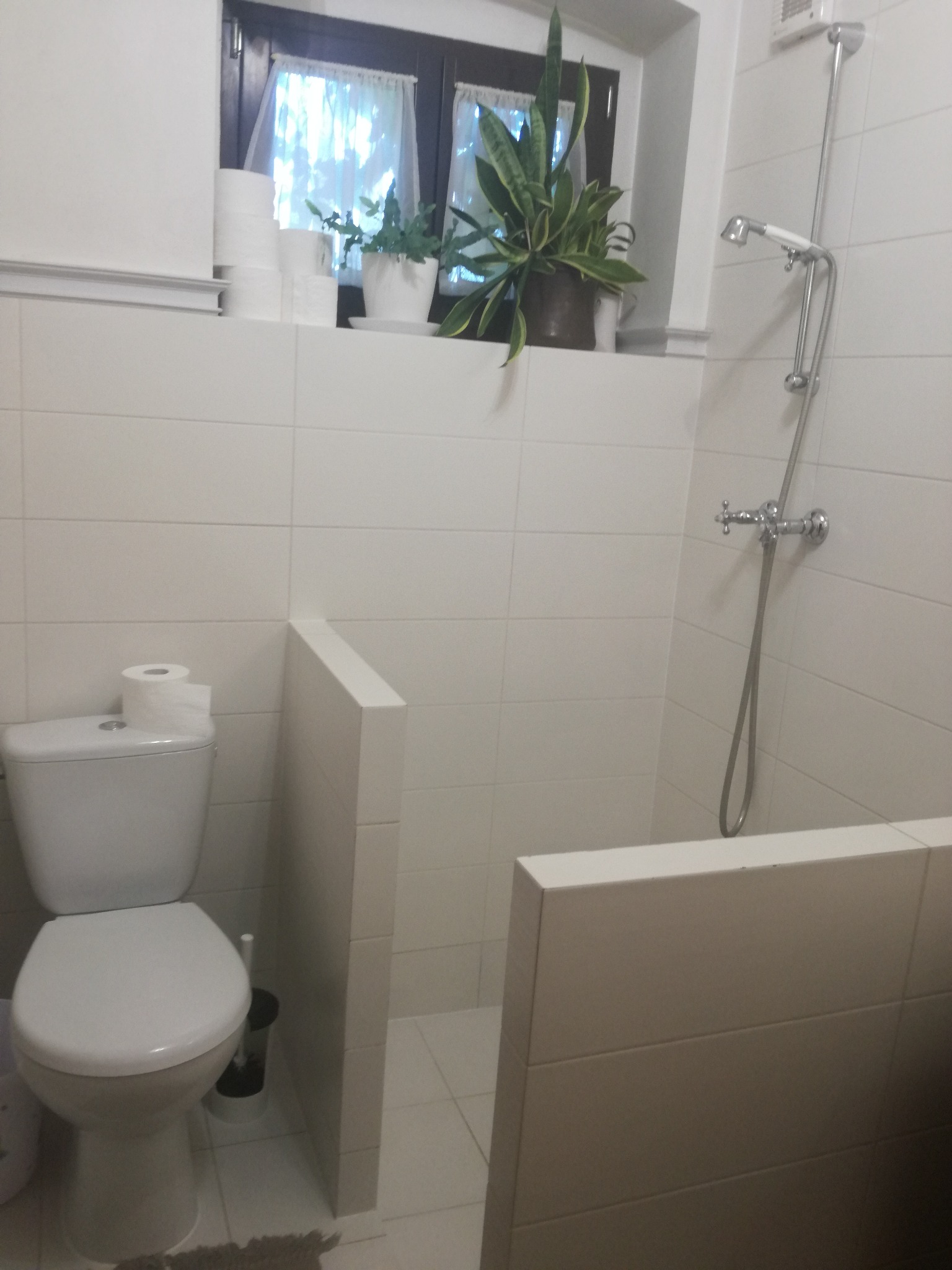 Jasna łazienka z białą glazurą, toaletą i kabiną prysznicową. Na parapecie okna widoczne rośliny doniczkowe i zapas papieru toaletowego.