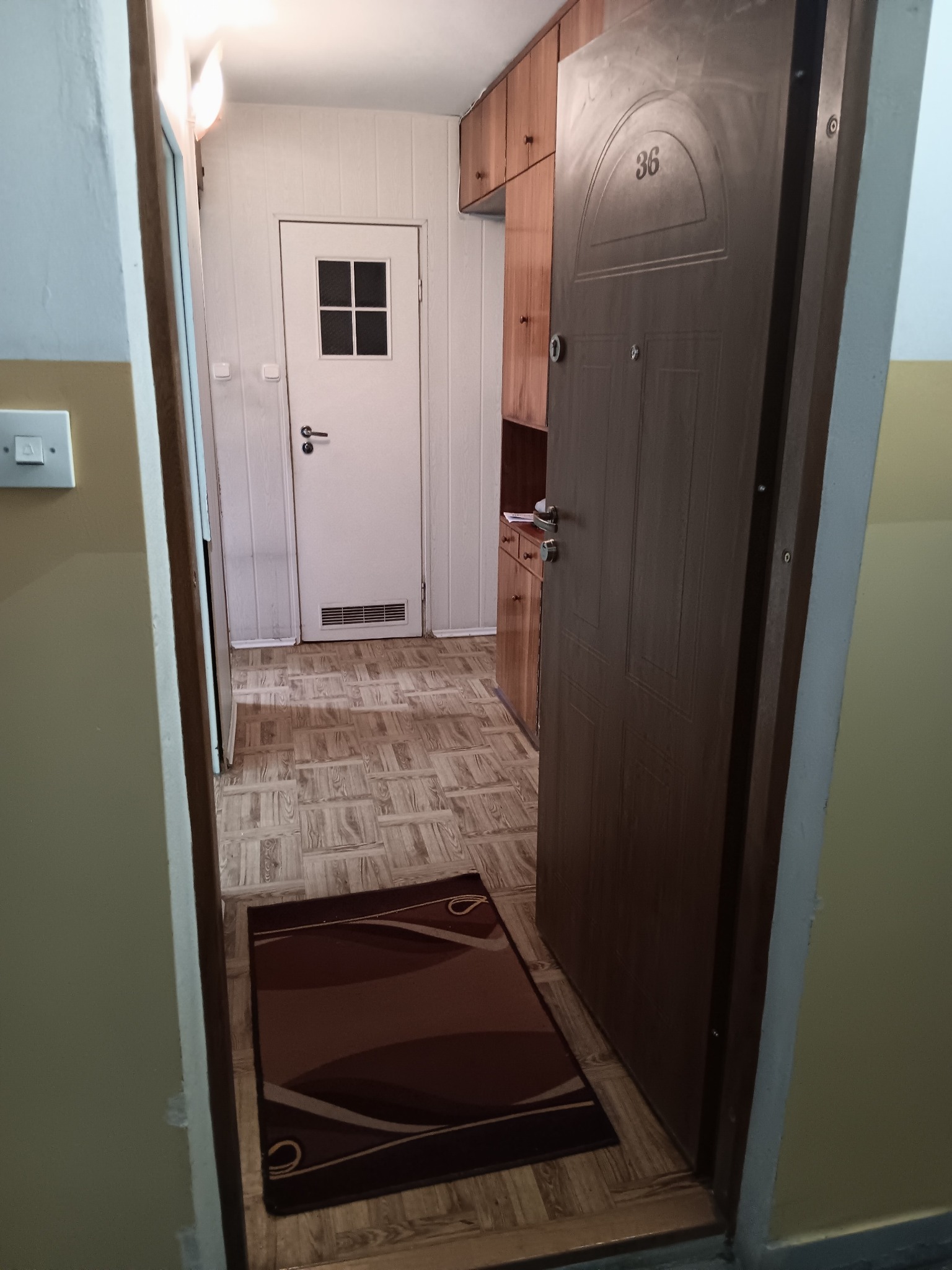 Otwarta, brązowa, antywłamaniowa  drzwi wejściowe z numerem 36, widok z korytarza na białe drzwi wewnętrzne z szybą i drewniane szafki. Na podłodze leży brązowy dywanik.