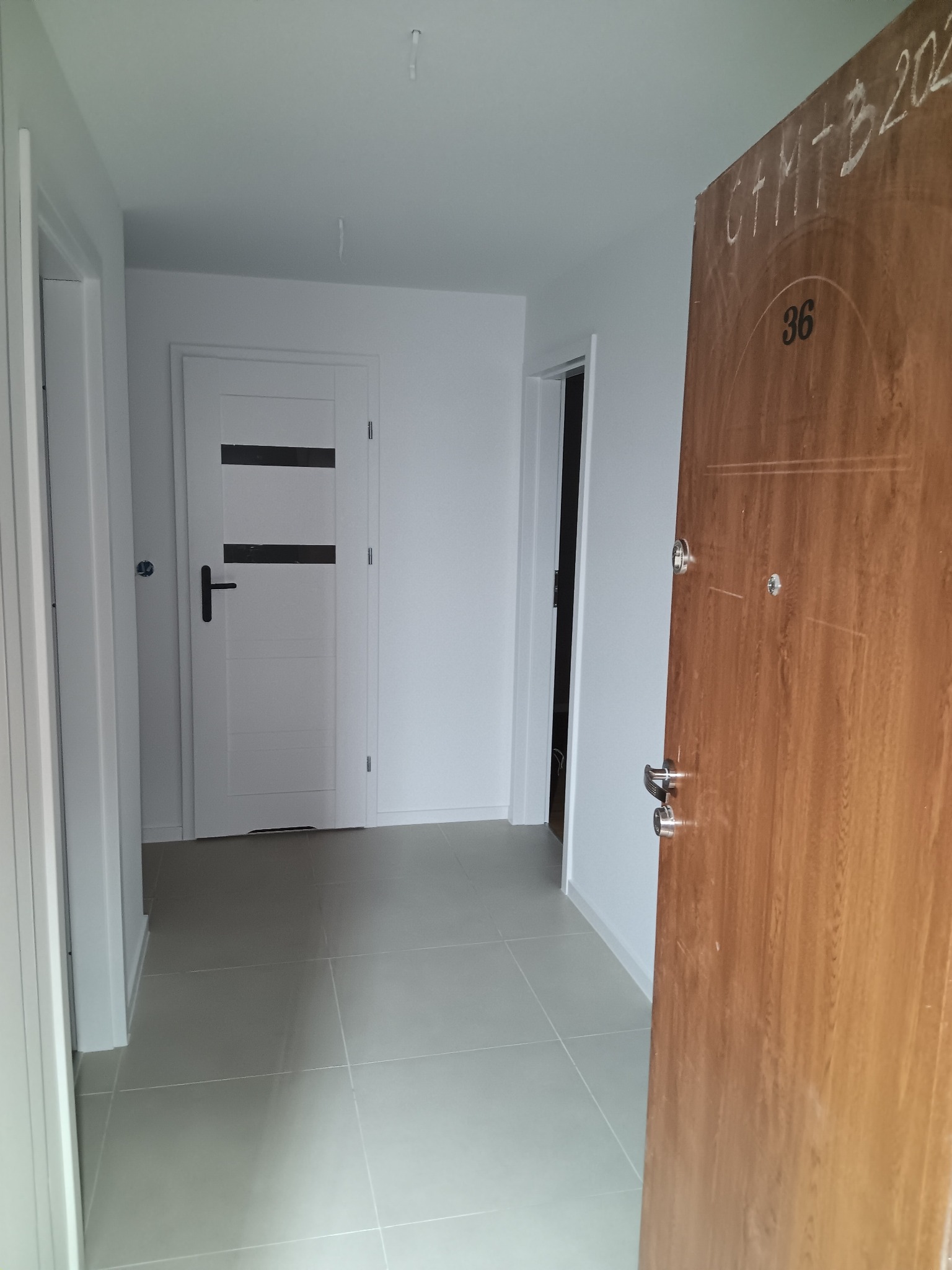 Wnętrze po remoncie: jasne ściany, szara podłoga z płytek, białe drzwi wewnętrzne z czarnymi akcentami i brązowe drzwi wejściowe z numerem 36.