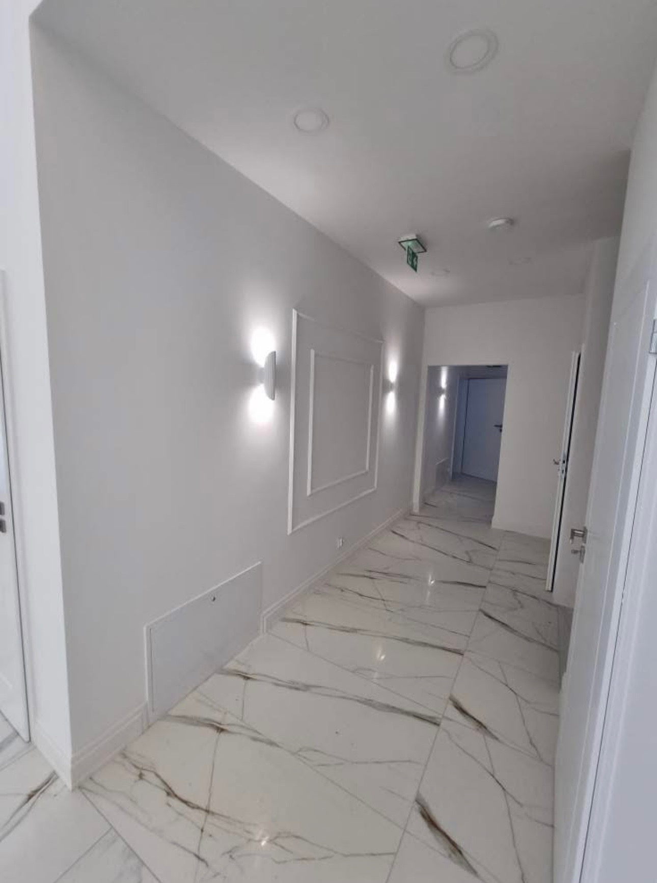 Biały korytarz z marmurową podłogą, oświetleniem ściennym i dekoracyjnymi ramami. Widok na dalszą część korytarza z drzwiami. Minimalistyczny design.