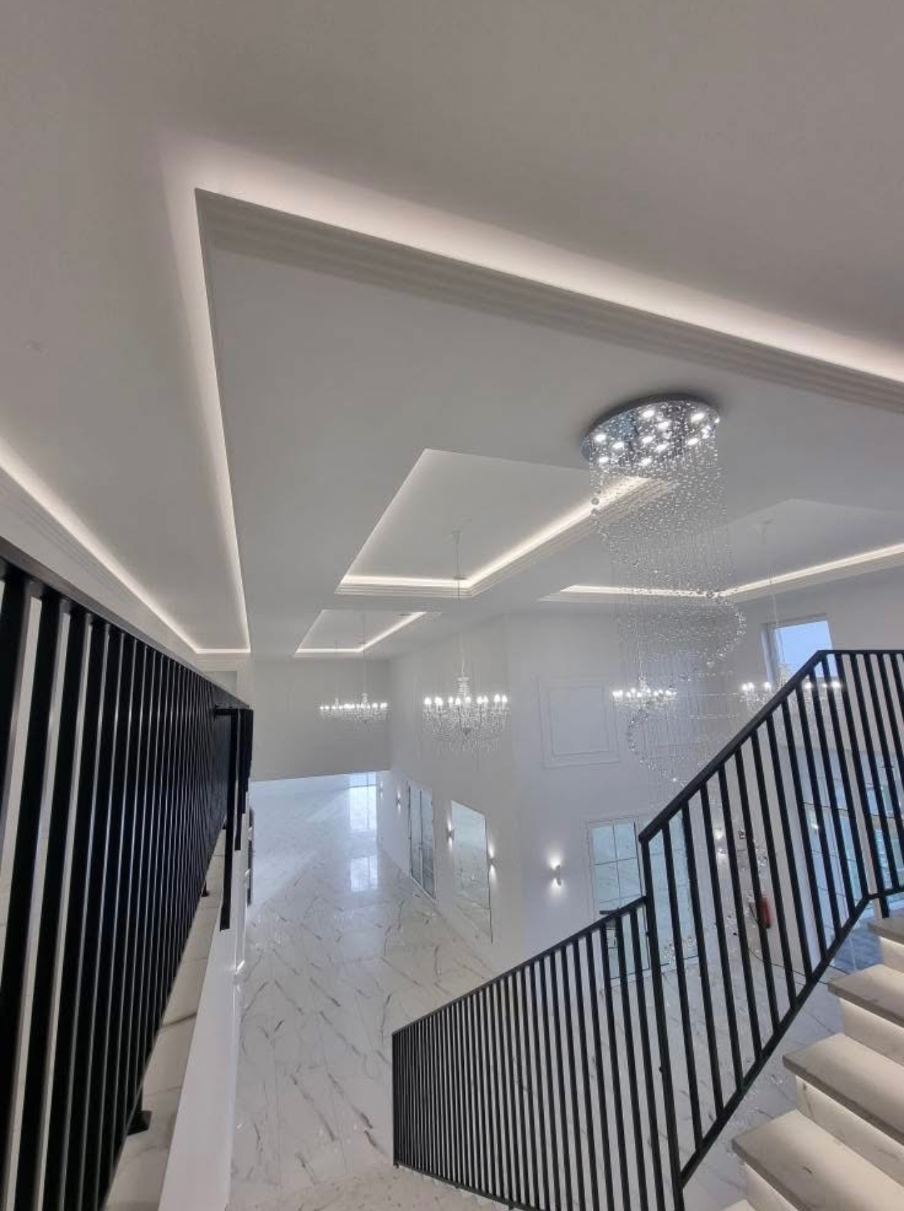 Nowoczesne wykończenie wnętrza: białe ściany, marmurowa podłoga i designerskie oświetlenie LED na suficie. Widok z góry na schody z czarną balustradą.