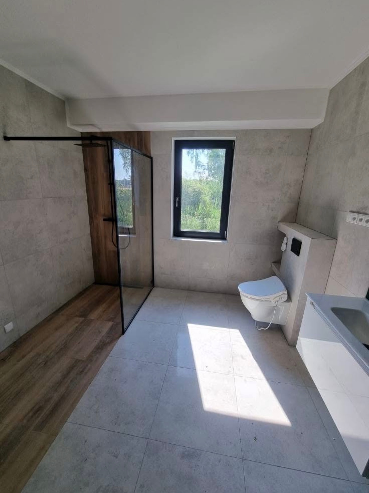 Nowoczesna łazienka z kabiną typu walk-in, wykończona płytkami w odcieniach szarości. Widoczne okno z widokiem na zieleń i minimalistyczna armatura.