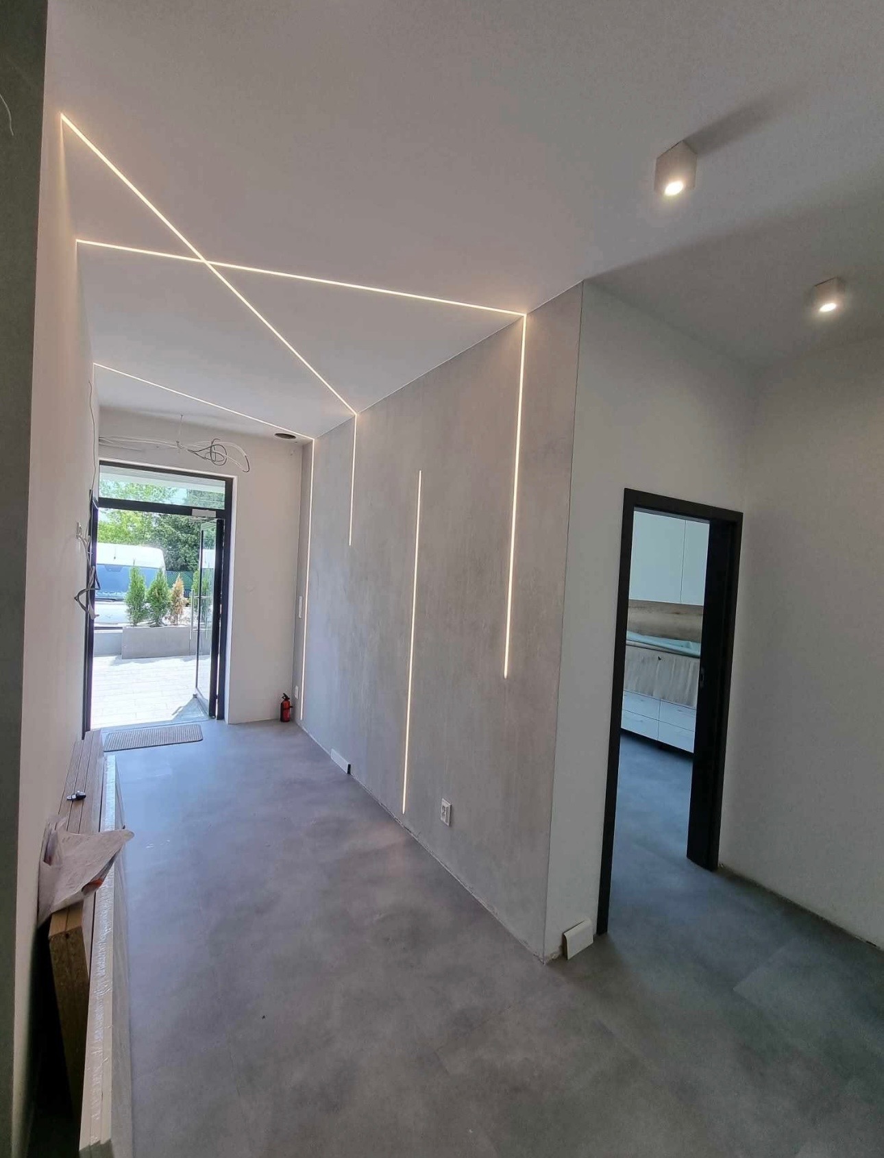 Nowoczesne oświetlenie LED w korytarzu z szarą posadzką i ścianami o surowym wykończeniu. Geometryczne linie światła na suficie i pionowe na ścianach.