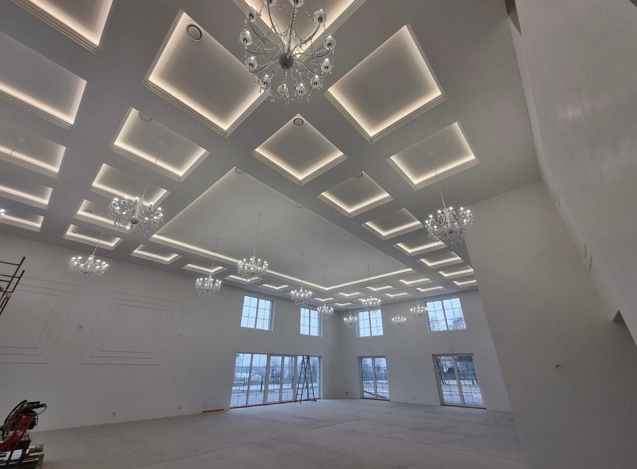 Sala balowa z białymi ścianami, oknami i dekoracyjnym oświetleniem sufitowym. Widoczne schody i sprzęt budowlany w rogu. Duża przestrzeń przygotowana do wykończenia.