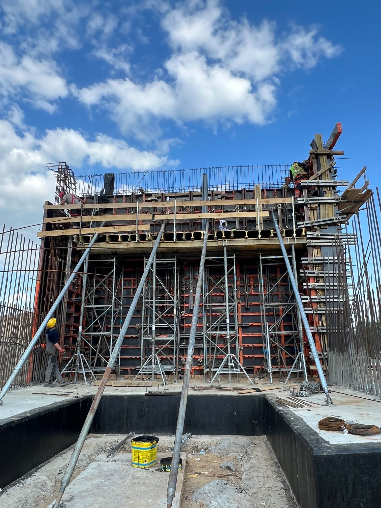 Budowa: Szalunek zbrojony na fundamencie, widoczni pracownicy w kaskach, stalowe podpory i niebieskie niebo z chmurami w tle. Prace budowlane w toku.