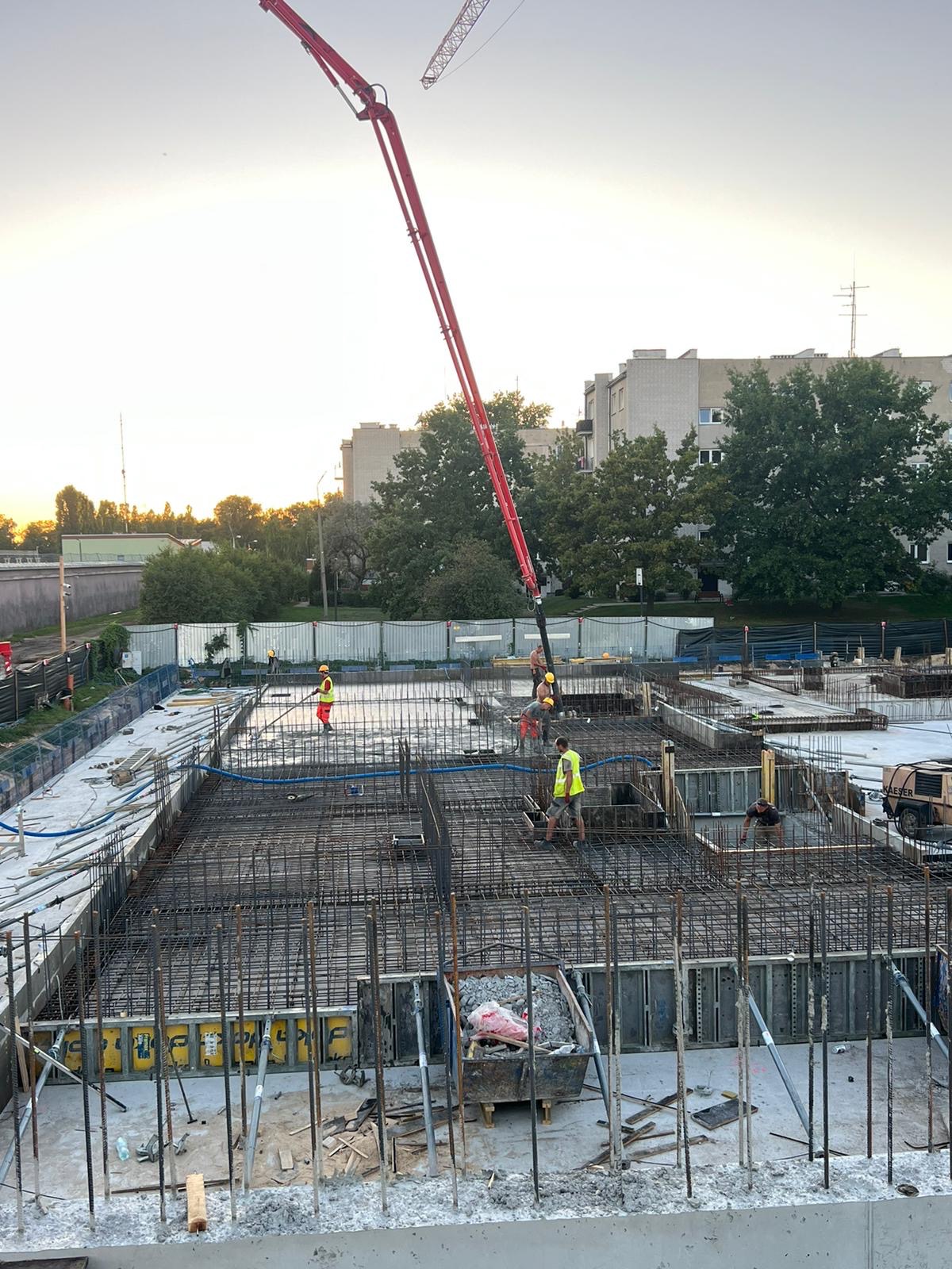 Wysoki kąt: robotnicy w kamizelkach wylewają beton na zbrojenie fundamentów budynku za pomocą pompy do betonu. W tle budynki mieszkalne i zachodzące słońce.