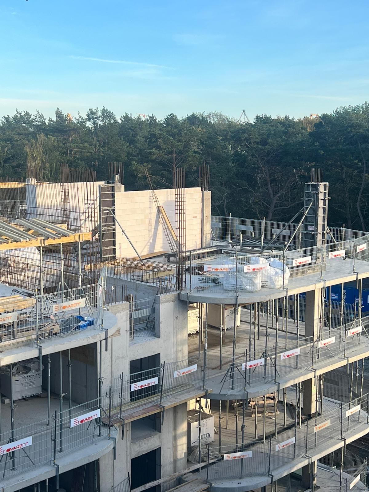 Wysoki budynek w trakcie budowy, widoczne szkielety ścian z betonu komórkowego, rusztowania i zbrojenia. W tle las i fragment nieba.