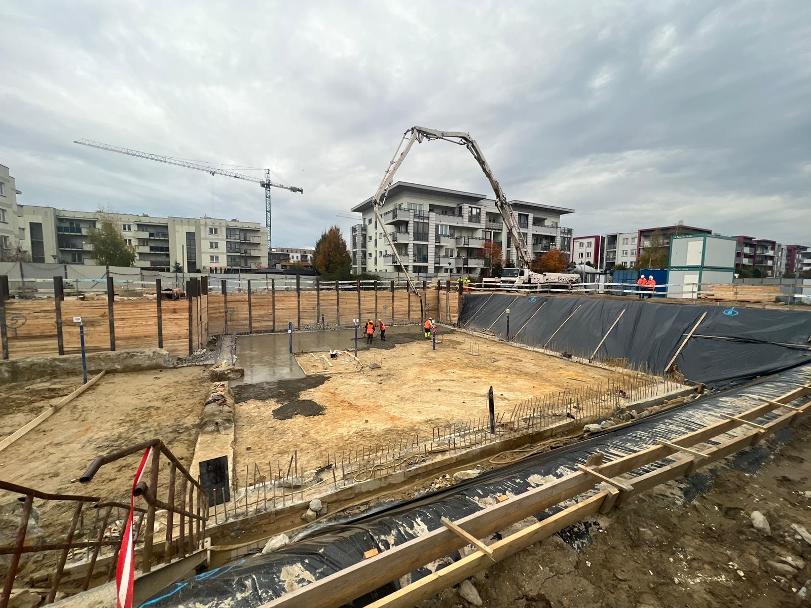 Wylewanie fundamentów pod budynek wielorodzinny. Na budowie pracownicy w pomarańczowych kamizelkach i betonowóz z wysięgnikiem. W tle budynki mieszkalne i żuraw.