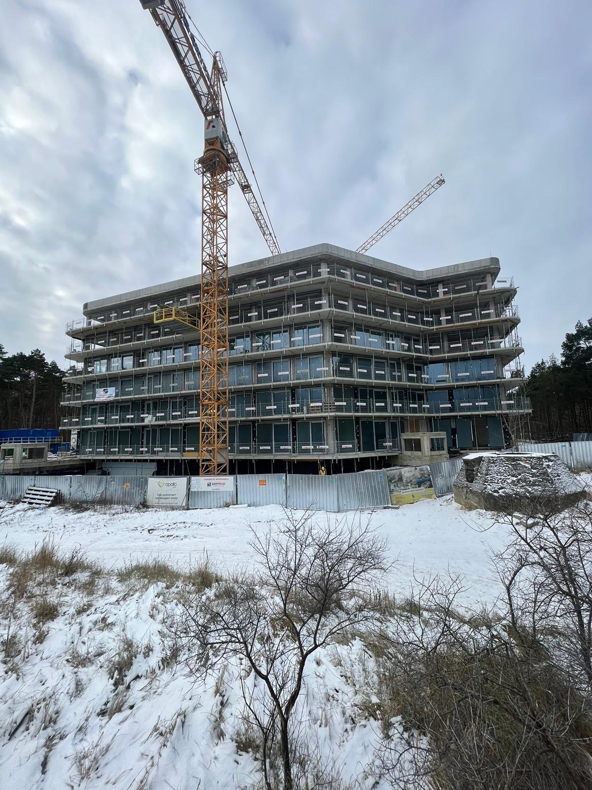 Wielopiętrowy budynek w stanie surowym zamkniętym, z widocznymi oknami i balkonami, otoczony ogrodzeniem i żurawiem budowlanym, zimowy krajobraz z śniegiem.
