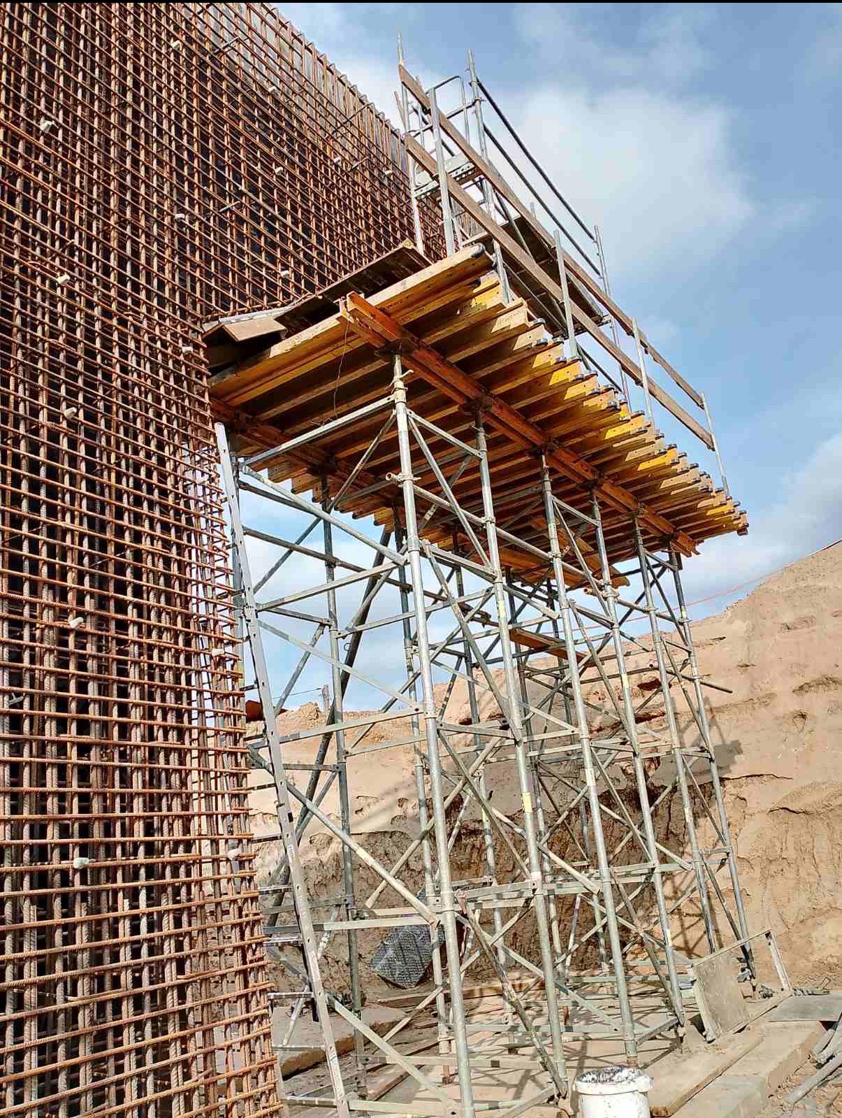Zbrojenie ściany z siatki stalowej i rusztowanie z deskowaniem, przygotowane do wylania betonu. Widok z dołu, niebo w tle.