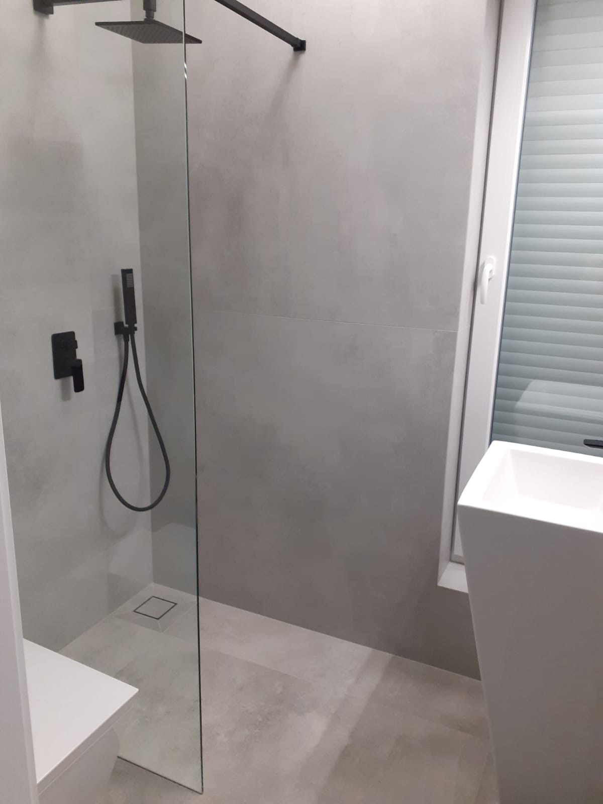 Minimalistyczna łazienka z kabiną typu walk-in, szara glazura, czarna armatura i biała umywalka. Widoczne detale wykończenia i nowoczesny design.