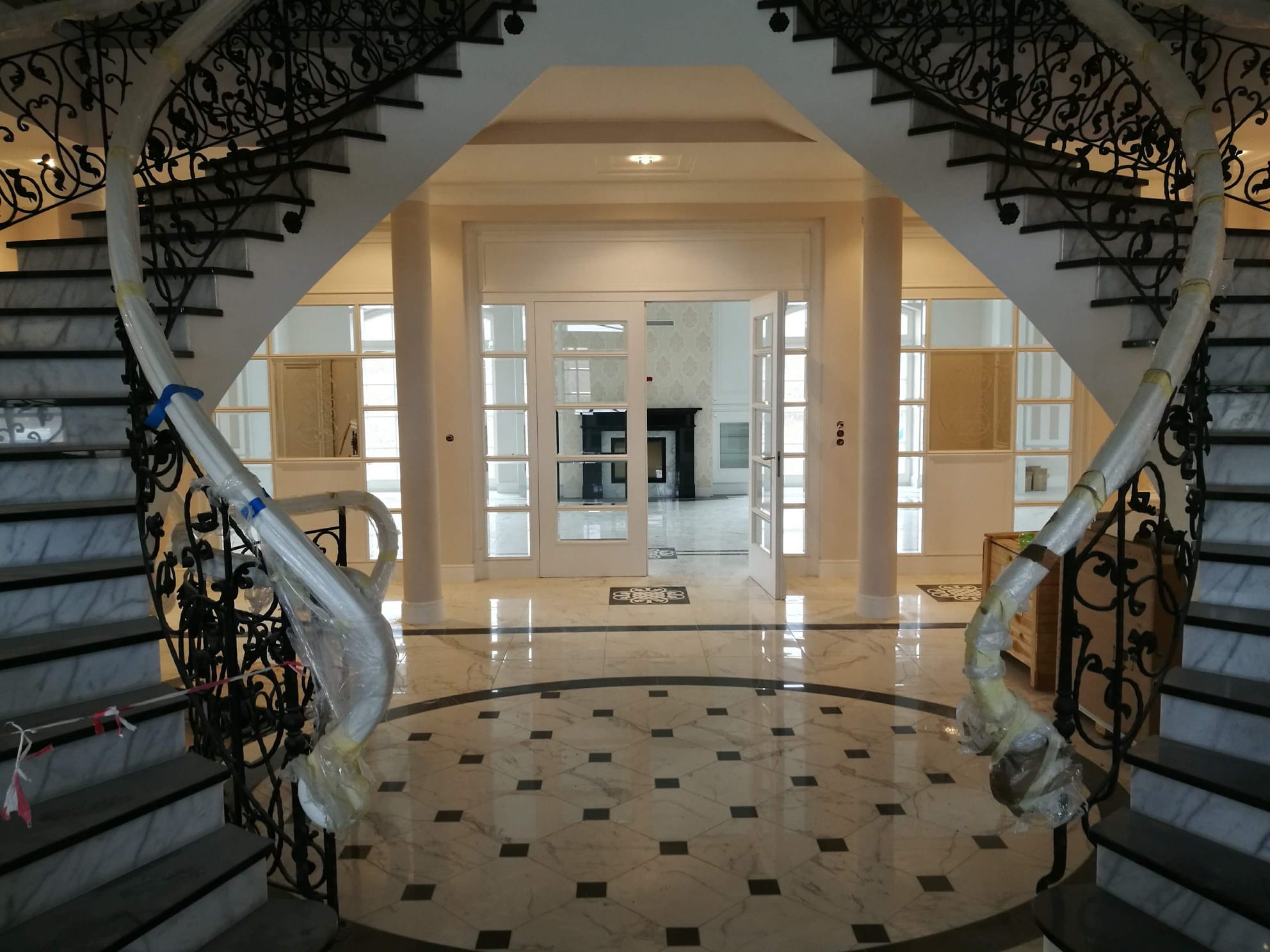 Elegancki hol z marmurową posadzką w geometryczny wzór i schodami z kutą balustradą, zabezpieczoną folią ochronną. Widok na otwarte, przeszklone drzwi.