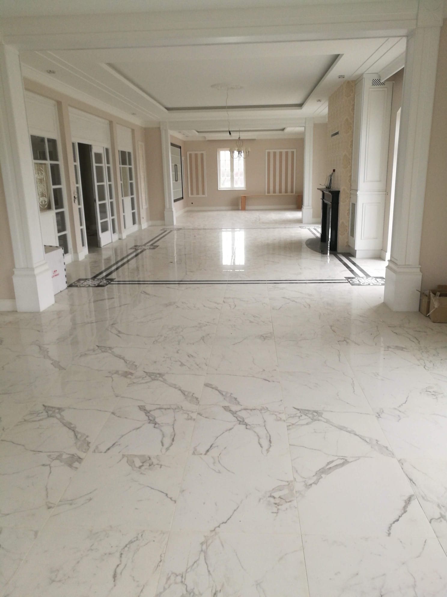 Jasne, marmurowe płytki podłogowe z dekoracyjnymi wstawkami w przestronnym salonie z kolumnami i kominkiem. Widok na dalszą część pomieszczenia z oknem.