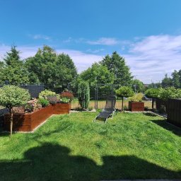 Ogrody po Naszymu - Zadbany ogród z równo przystrzyżonym trawnikiem, drewnianymi donicami z roślinami i leżakiem. Widoczne drzewa i krzewy w tle, ogrodzenie panelowe.