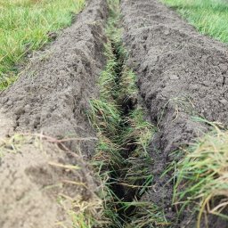 Ogrody po Naszymu - Wykopany rów w ziemi, przygotowany pod trawnik z rolki. Widoczne resztki trawy w rowie i trawa po bokach. Perspektywa z dołu, ziemia w odcieniach szarości i brązu.