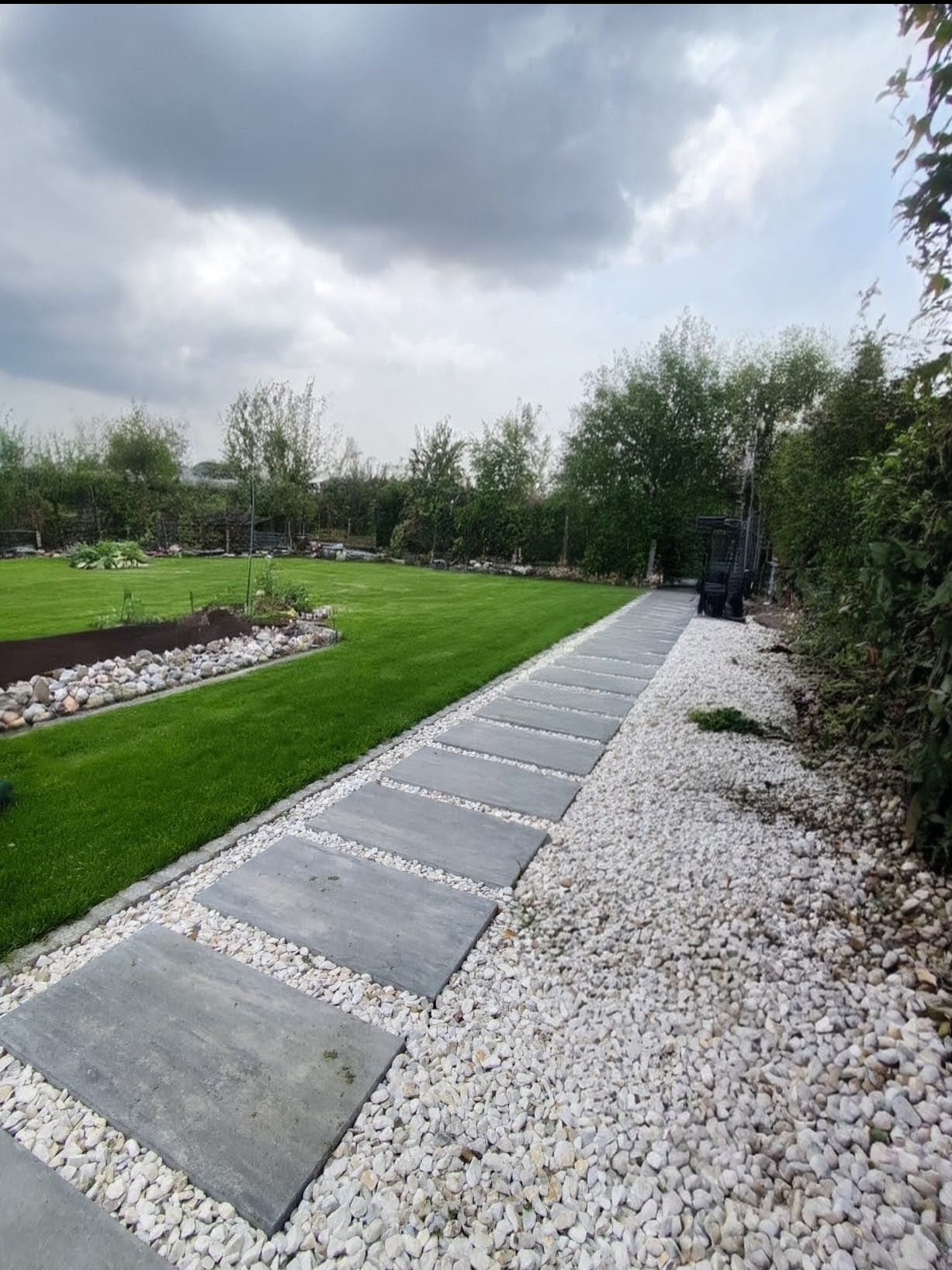 Ogród z trawnikiem i ścieżką z szarych płyt oddzielonych białym żwirem. W tle drzewa i krzewy pod pochmurnym niebem. Widoczny fragment ogrodu z kamieniami.
