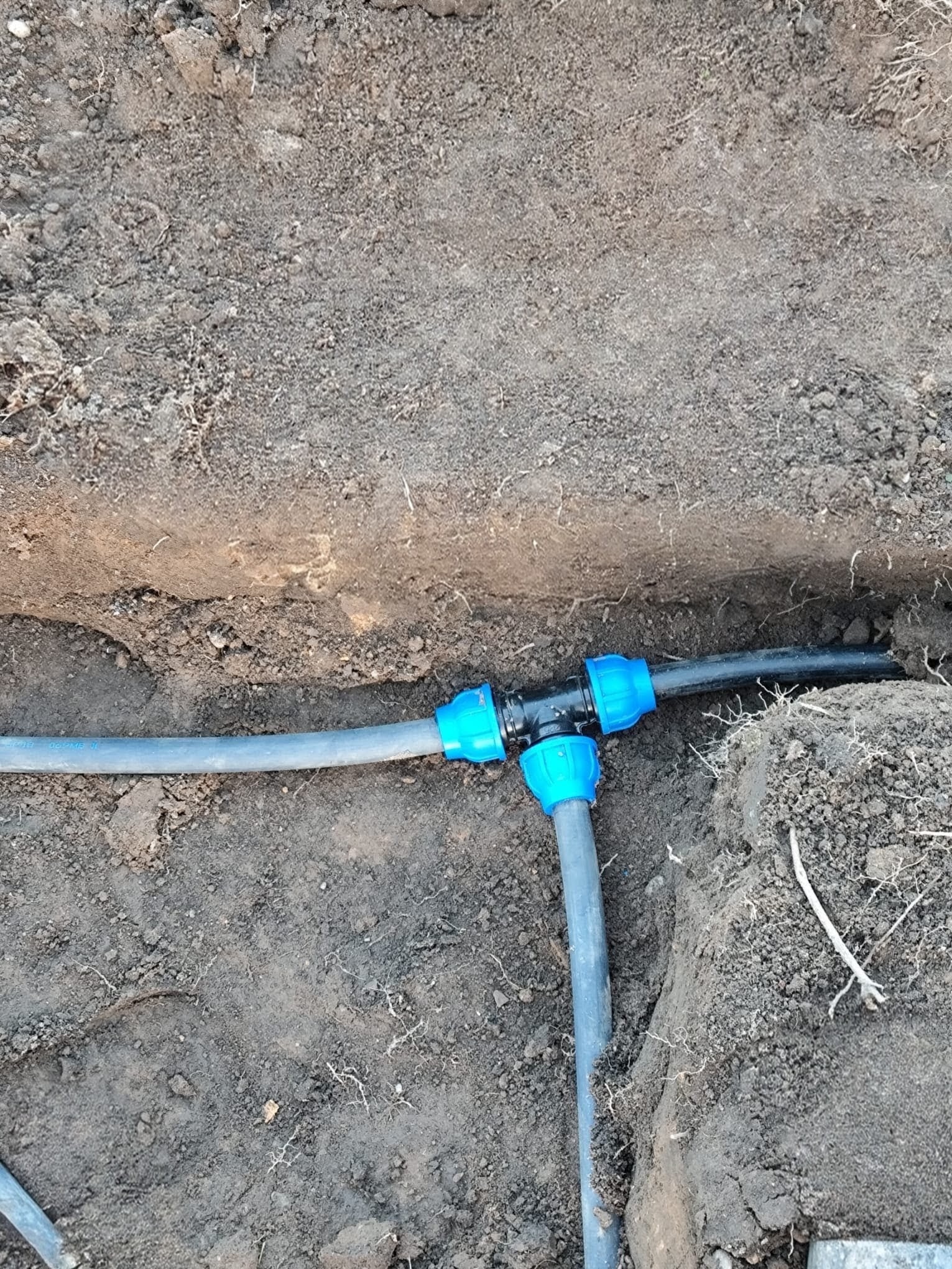Połączenie rur z niebieskimi złączkami w wykopie ziemnym. Widoczne fragmenty rur i ziemia. Instalacja podziemna w trakcie montażu.