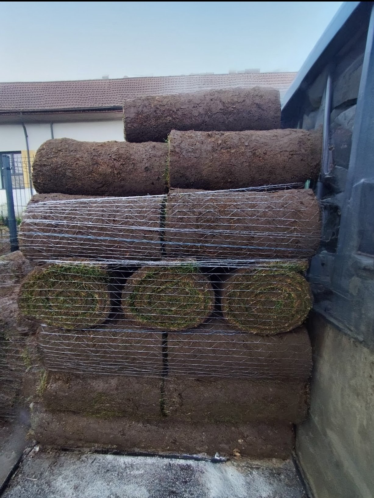 Stos rolek trawy w siatce, ułożonych w skrzyni ładunkowej pojazdu. Widoczne warstwy darni i ziemi, zarys budynku w tle. Gotowe do transportu.
