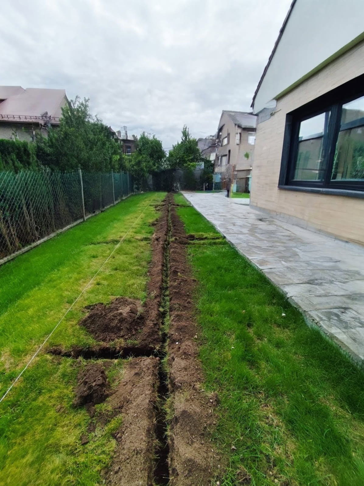 Wykopany wąski rów w trawniku, prawdopodobnie pod instalację nawadniającą lub drenaż. Widoczne pryzmy ziemi. W tle budynki mieszkalne i ogrodzenie.