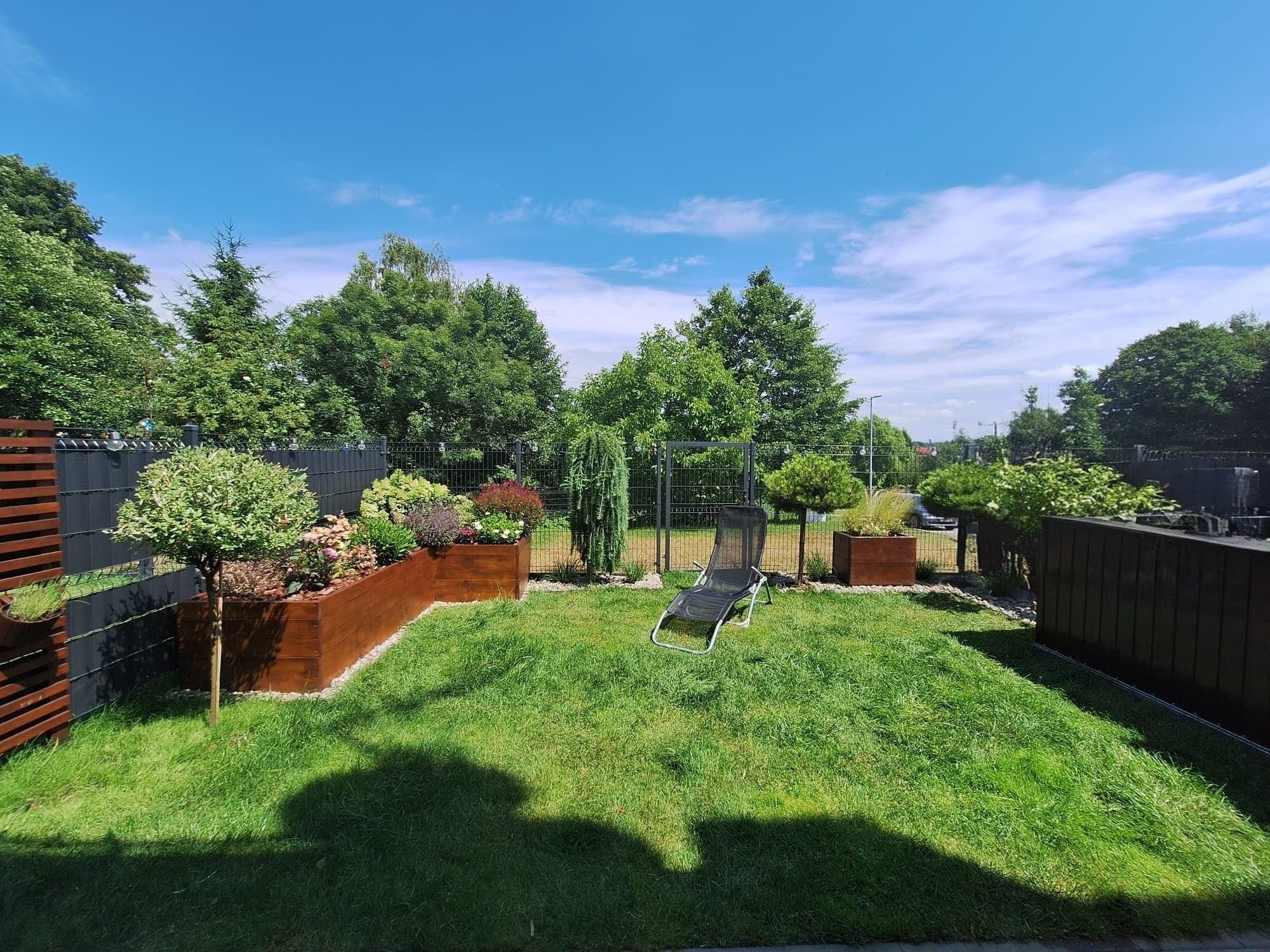 Zadbany ogród z równo przystrzyżonym trawnikiem, drewnianymi donicami z roślinami i leżakiem. Widoczne drzewa i krzewy w tle, ogrodzenie panelowe.