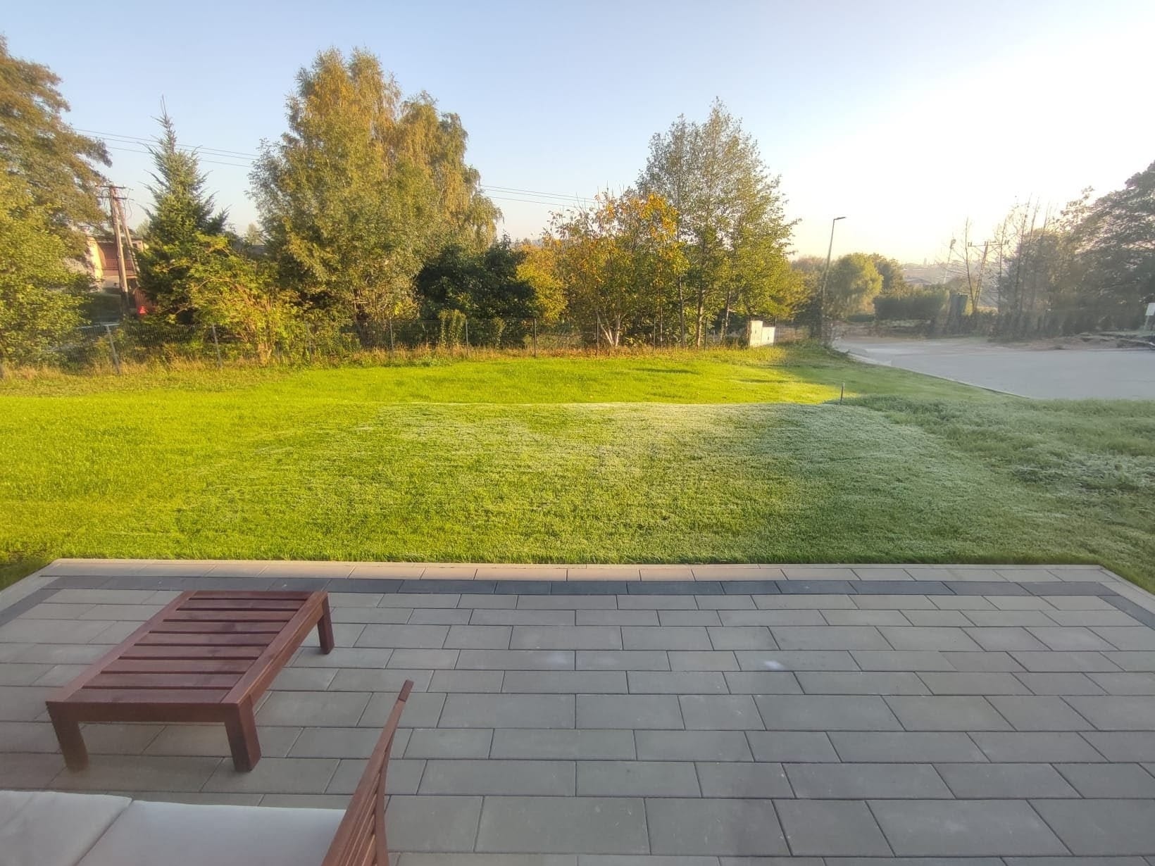 Poranna rosa na trawniku widziana z tarasu z kostki brukowej. W tle drzewa i krzewy w jesiennej kolorystyce. Ujęcie z niskiej perspektywy.