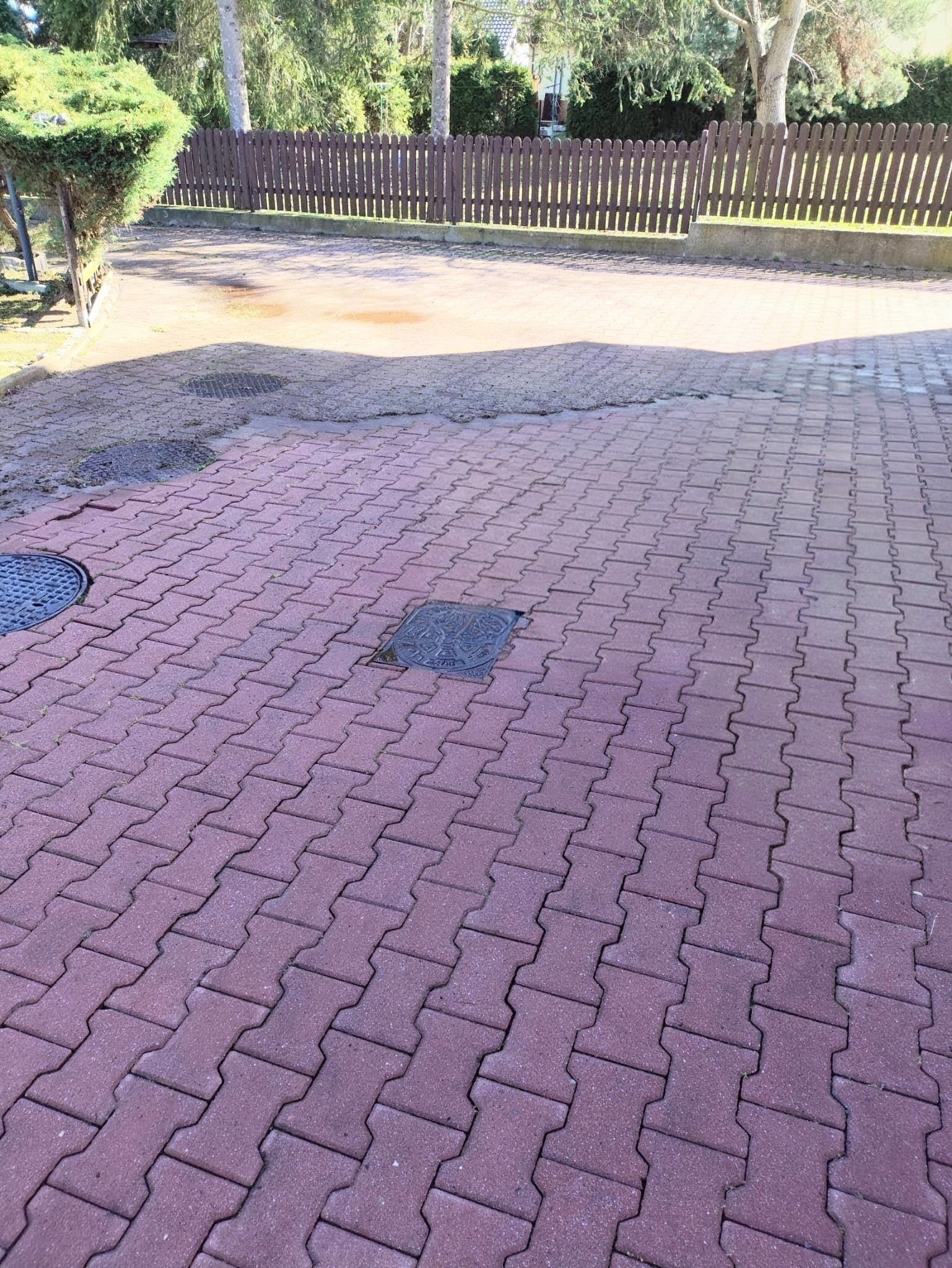 Ułożona, czerwona kostka brukowa w kształcie litery H, z widocznymi studzienkami kanalizacyjnymi. W tle drewniany płot i zieleń.