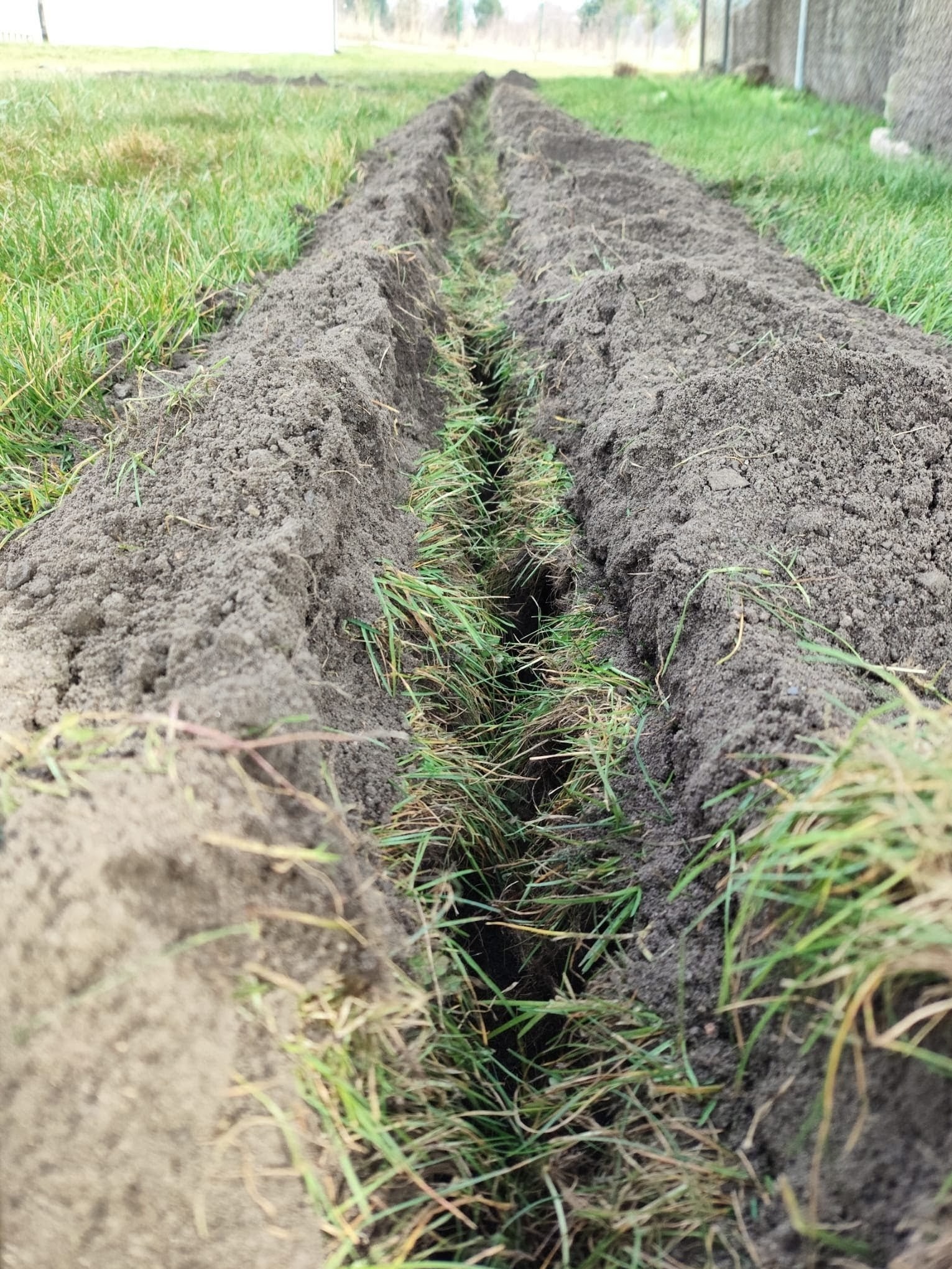 Wykopany rów w ziemi, przygotowany pod trawnik z rolki. Widoczne resztki trawy w rowie i trawa po bokach. Perspektywa z dołu, ziemia w odcieniach szarości i brązu.