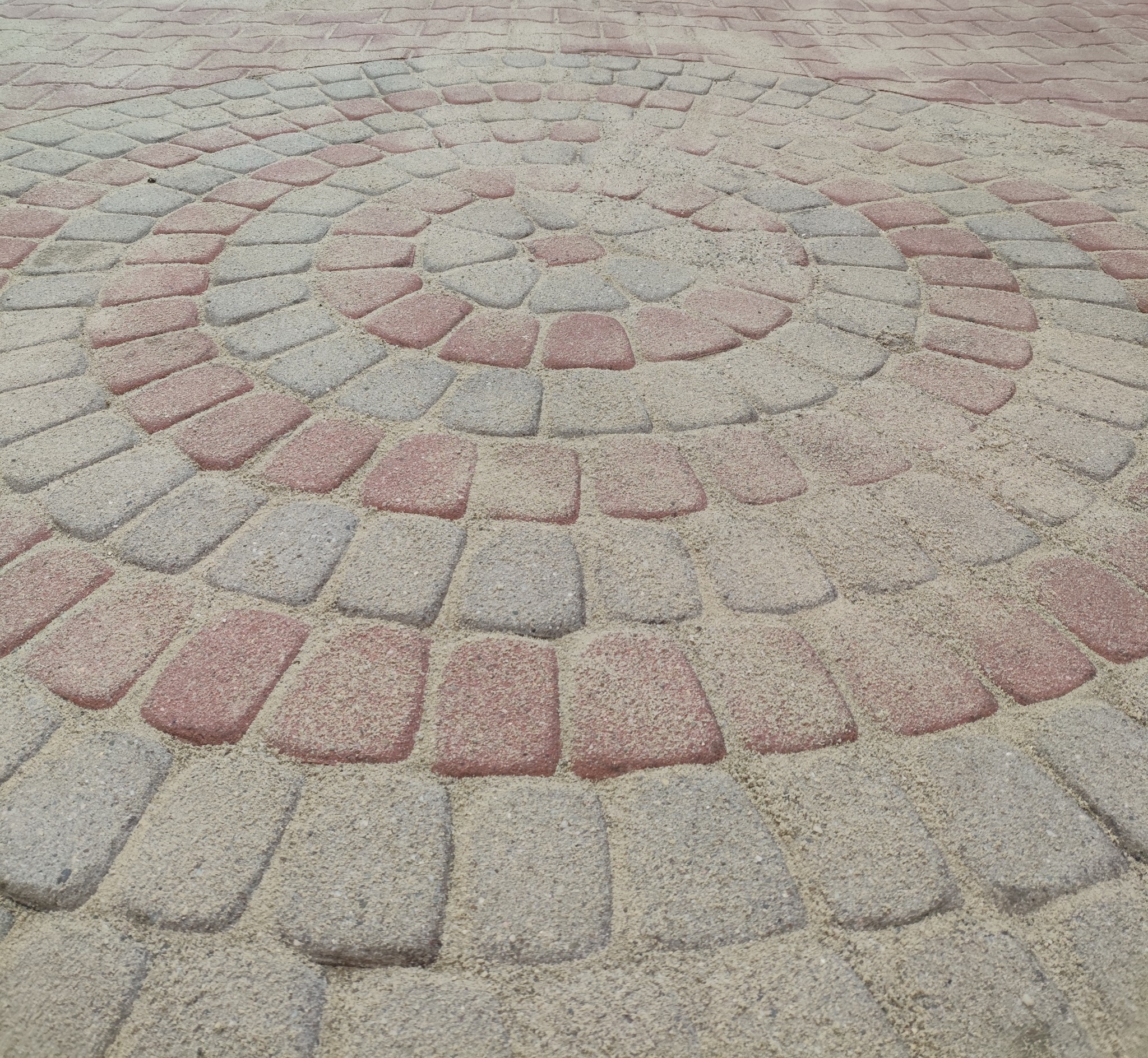 Ułożona kostka brukowa w okrągły wzór z naprzemiennymi rzędami szarych i czerwonych kostek. Widok z góry, perspektywa z dołu. Piasek między kostkami.