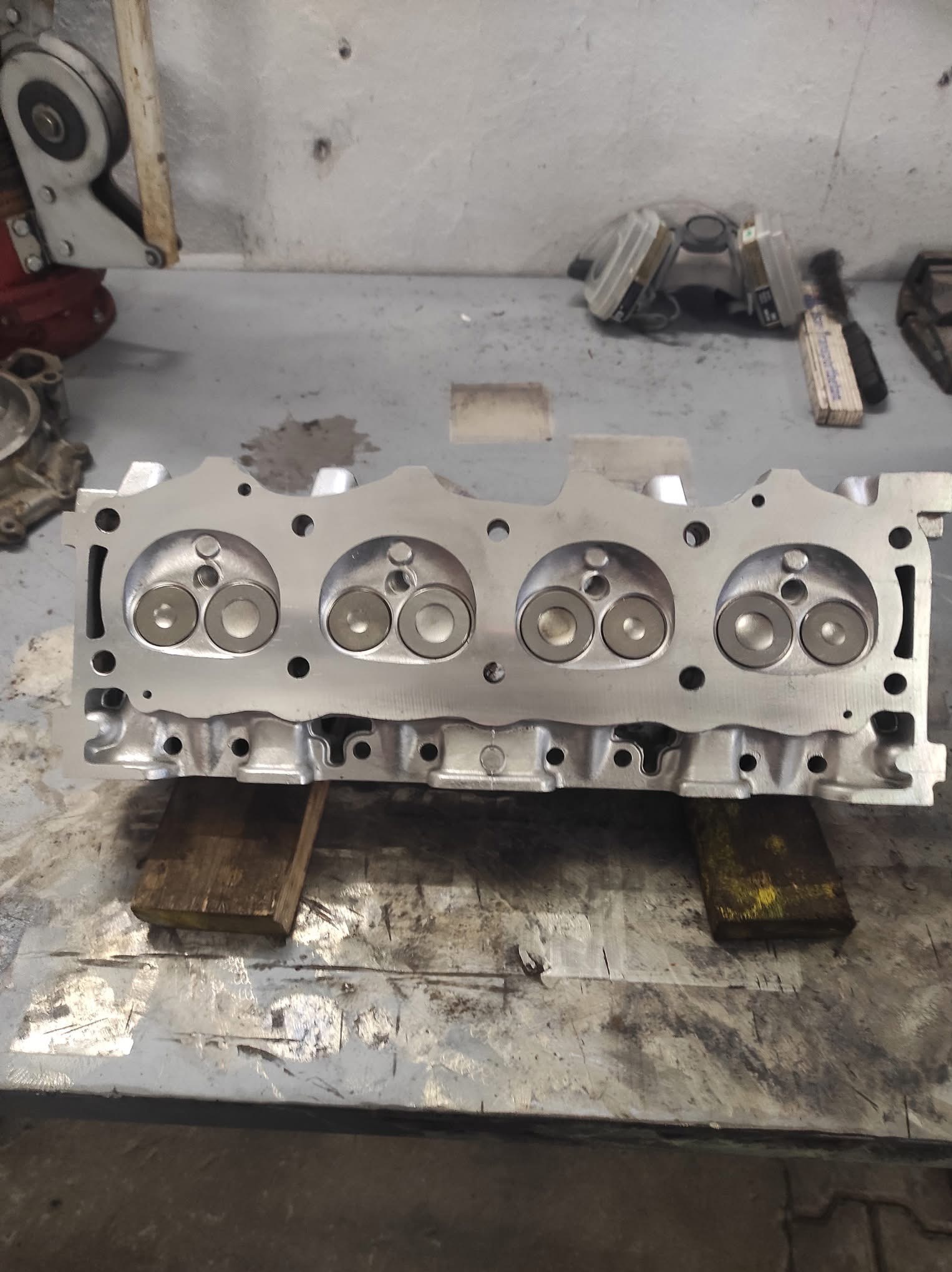 Oczyszczona głowica silnika spalinowego, cztery cylindry, widoczne zawory i kanały, leżąca na drewnianych podkładkach na warsztatowym stole.