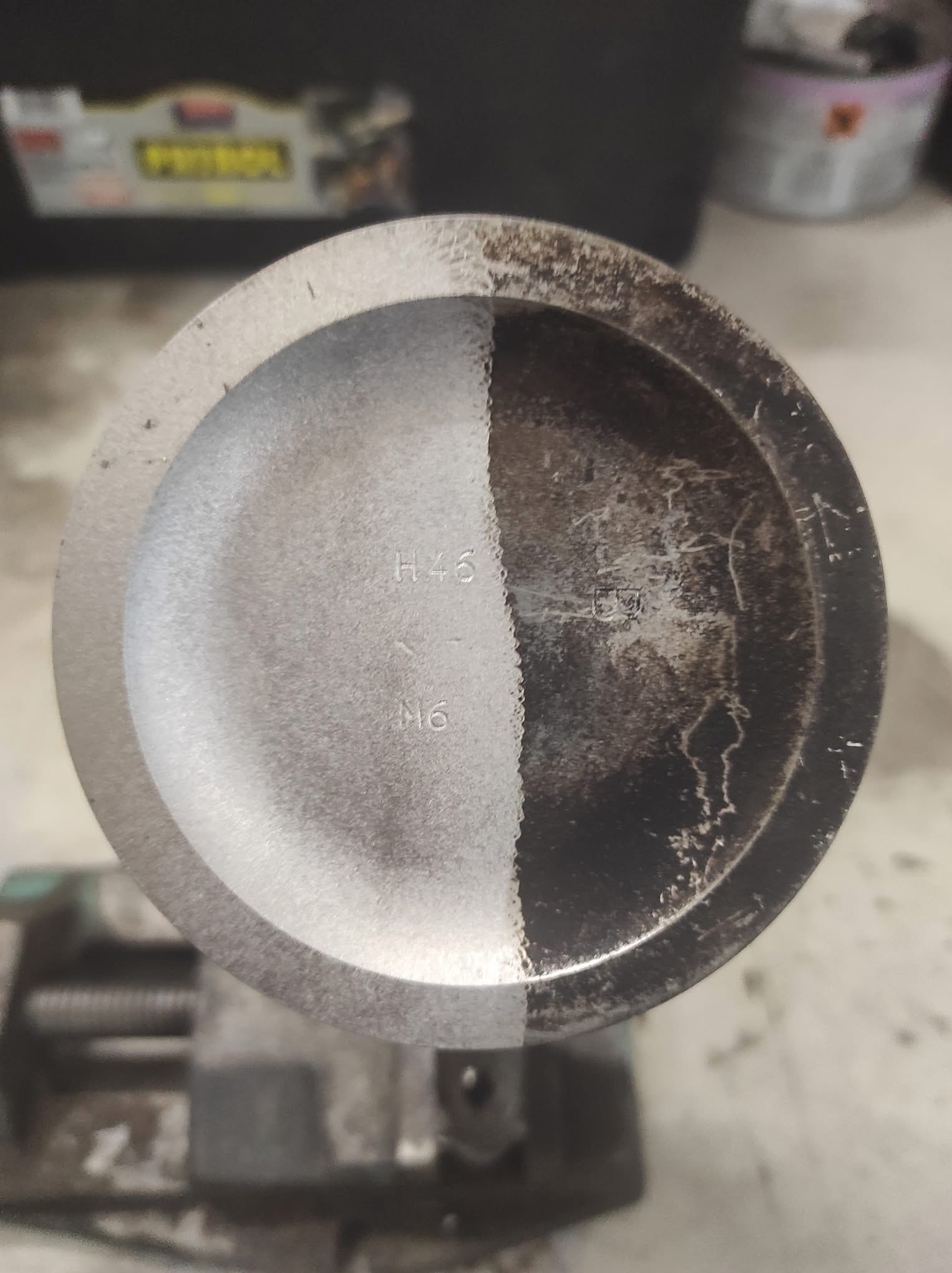 Częściowo wyczyszczony tłok silnika w imadle, prezentujący efekt przed i po czyszczeniu. Lewa strona tłoka jest czysta, prawa pokryta nagarem.