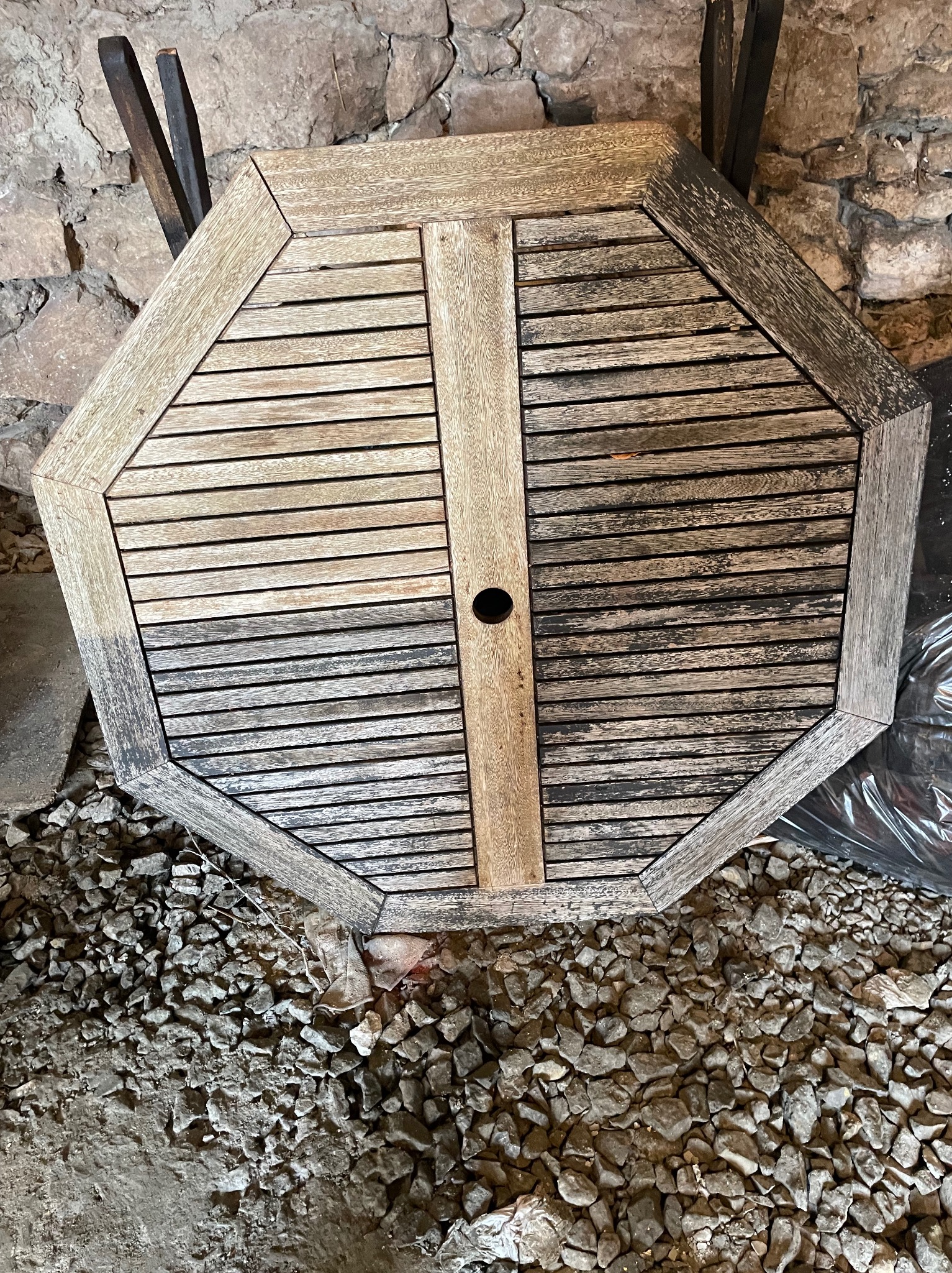 Ośmiokątny, drewniany blat stołu z widoczną różnicą w czystości połowy powierzchni, leżący na kamienistym podłożu, na tle kamiennej ściany.