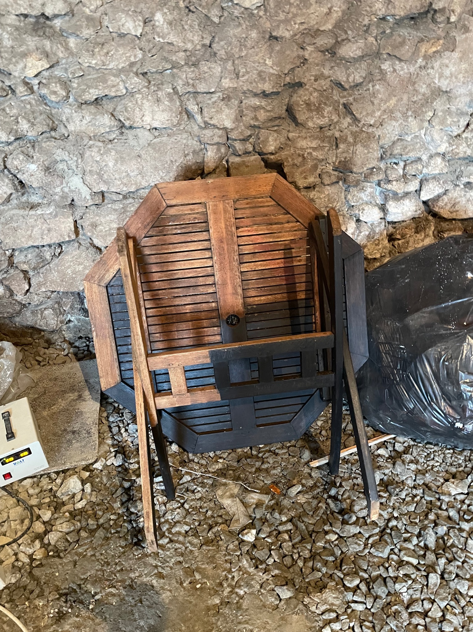 Drewniany, ośmiokątny stół z krzesłami w trakcie renowacji, częściowo pomalowany na czarno, oparty o kamienną ścianę w piwnicy.