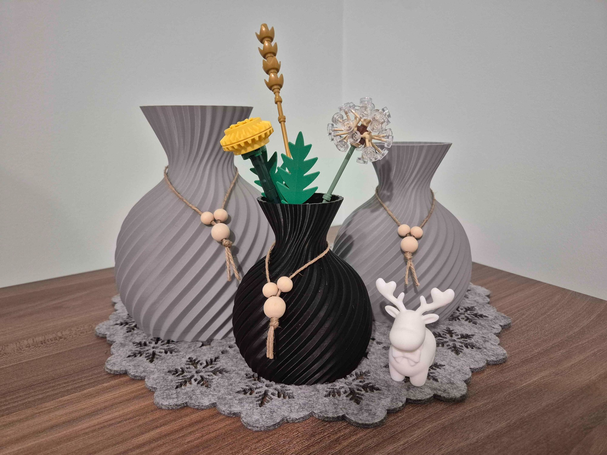 Trzy wazony z druku 3D (dwa szare, jeden czarny) z dekoracjami na ażurowej serwetce. Biały renifer z druku 3D. Widok z bliska, neutralne oświetlenie.