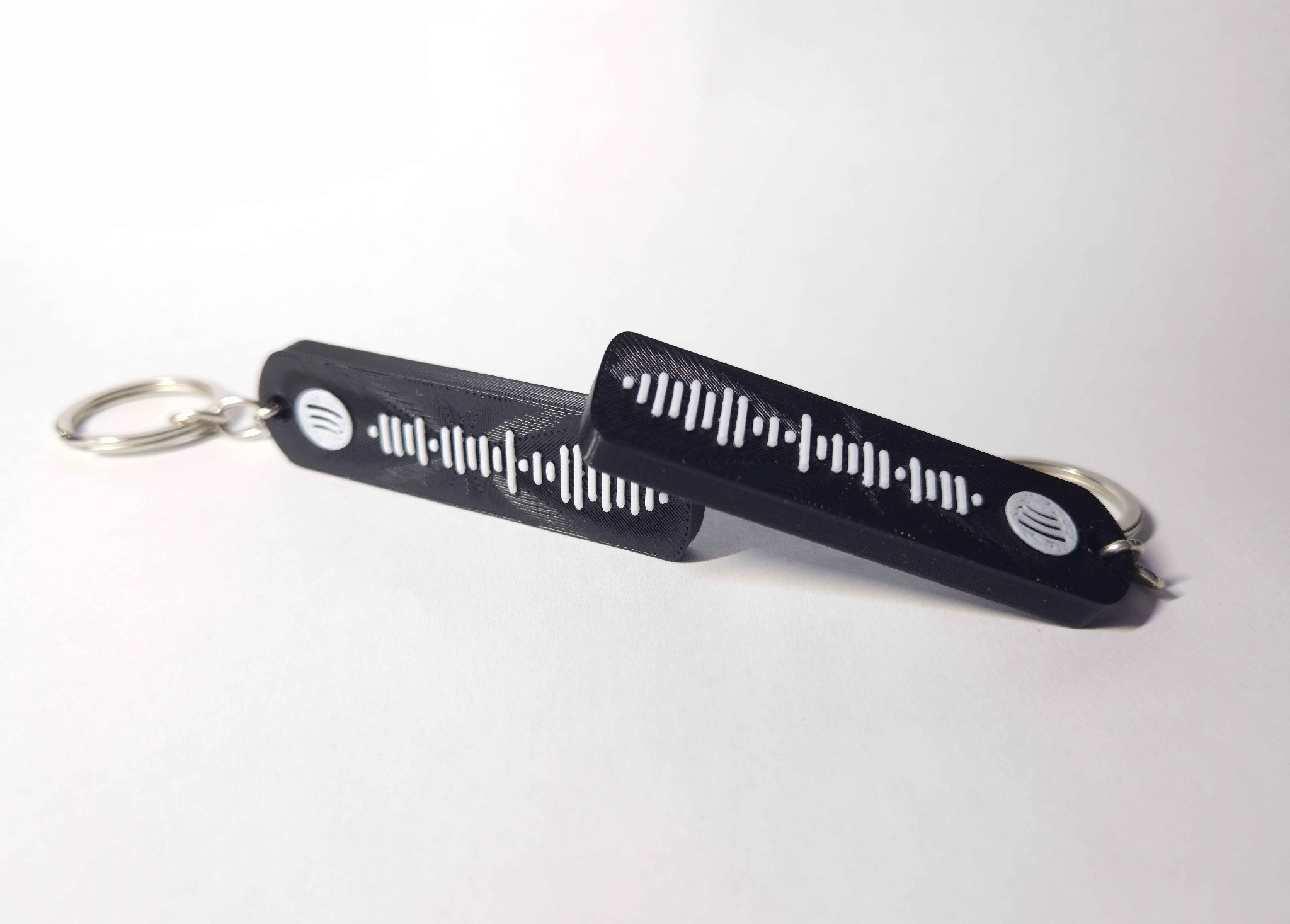 Dwa czarne breloczki z kodem Spotify, wydrukowane w 3D, leżą na białym tle. Widoczne drobne nierówności powierzchni charakterystyczne dla druku 3D.