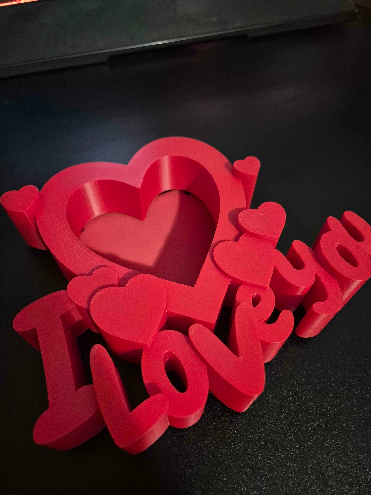 Czerwony, przestrzenny napis 'I Love You' z sercami, wydrukowany w 3D. Widoczna faktura druku, leży na czarnym, matowym tle. Ujęcie z bliska, z góry.