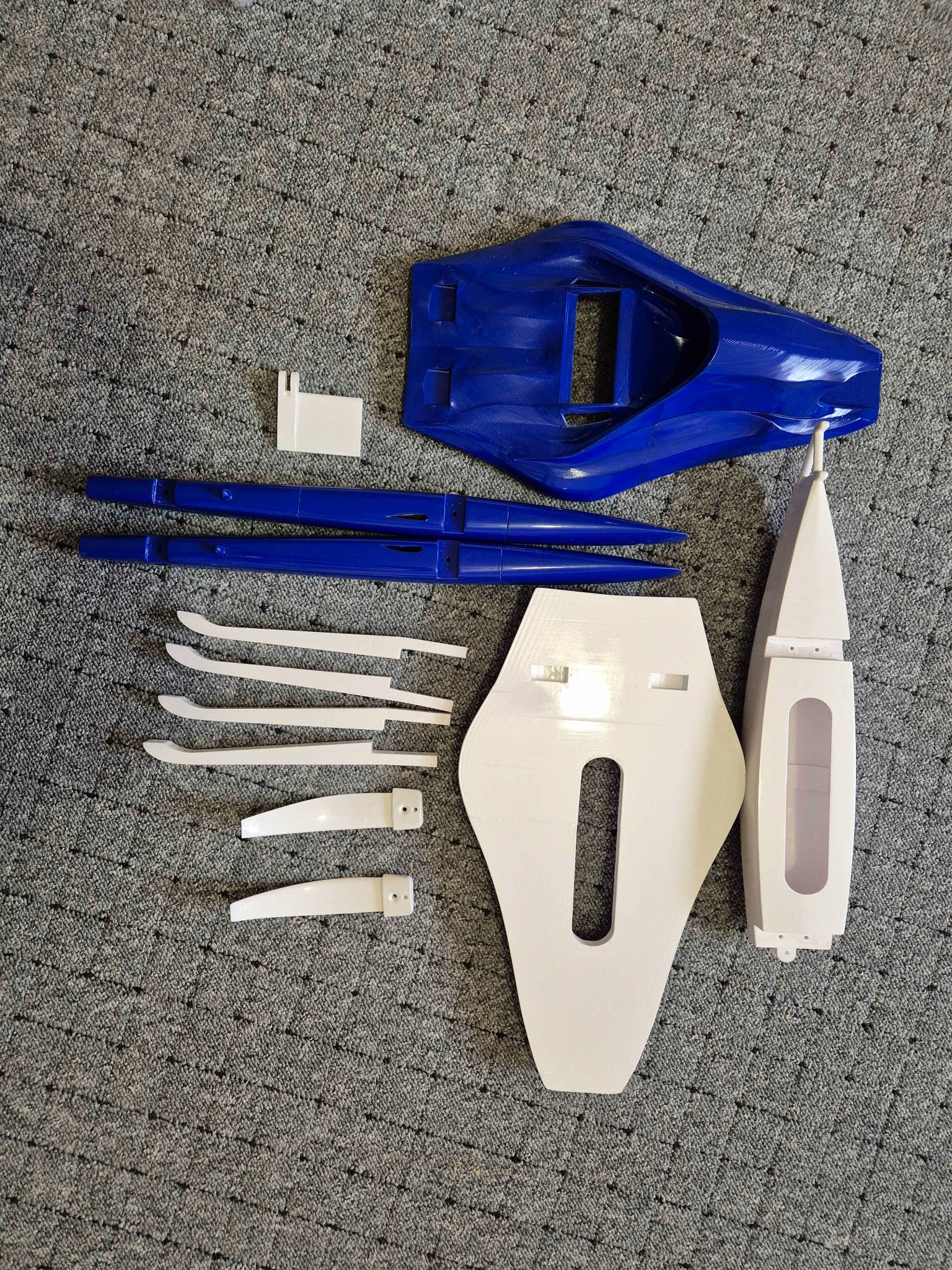 Elementy modelu trimaranu, wydrukowane w 3D, w kolorach białym i niebieskim, rozłożone na szarym tle. Widoczne ślady warstw wydruku.