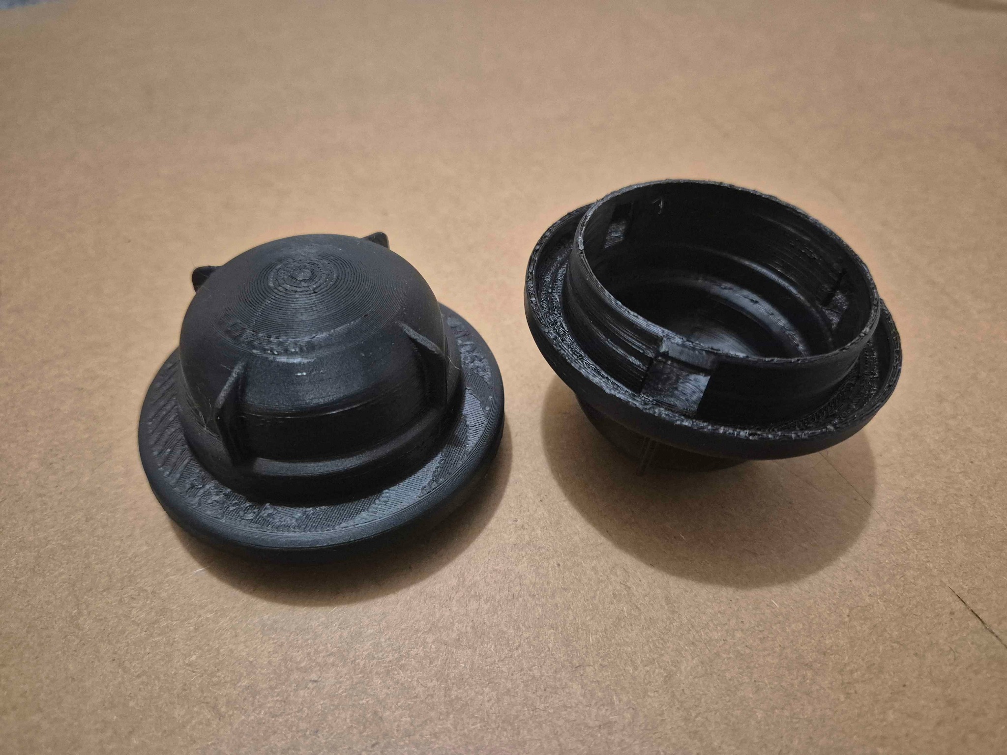 Czarne, rozdzielone elementy z tworzywa sztucznego, wydrukowane w technologii 3D, leżące na beżowym tle. Widoczne warstwy druku i detale konstrukcyjne.