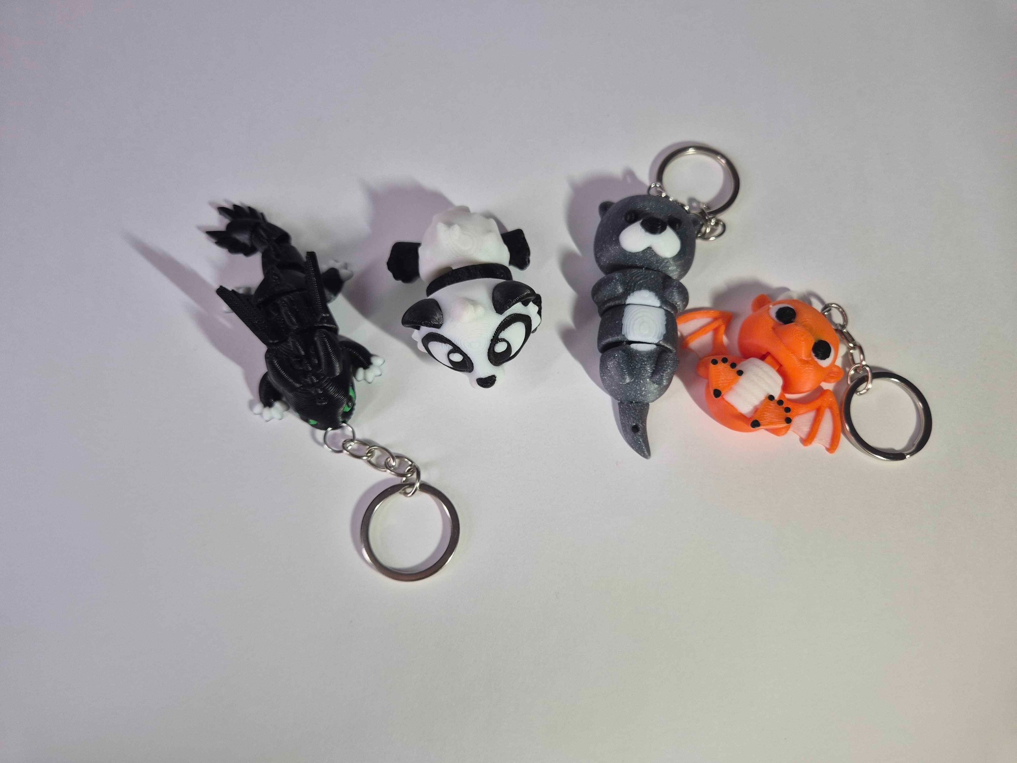 Cztery breloczki z motywami zwierzęcymi, wykonane w technologii druku 3D, leżą na białym tle. Widoczny smok, panda, wydra i pomarańczowy stworek.