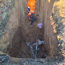 DRILL EXPERT Wojciech Świątek - Pracownik w pomarańczowym ubraniu w głębokim wykopie z rurą. Widoczny kubeł i narzędzia. Przewiert sterowany w trakcie realizacji.