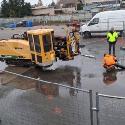 DRILL EXPERT Wojciech Świątek - Żółta maszyna Vermeer D24x40II do wiercenia horyzontalnego na mokrym placu budowy, pracownik w pomarańczowej kurtce łączy rury, drugi nadzoruje.