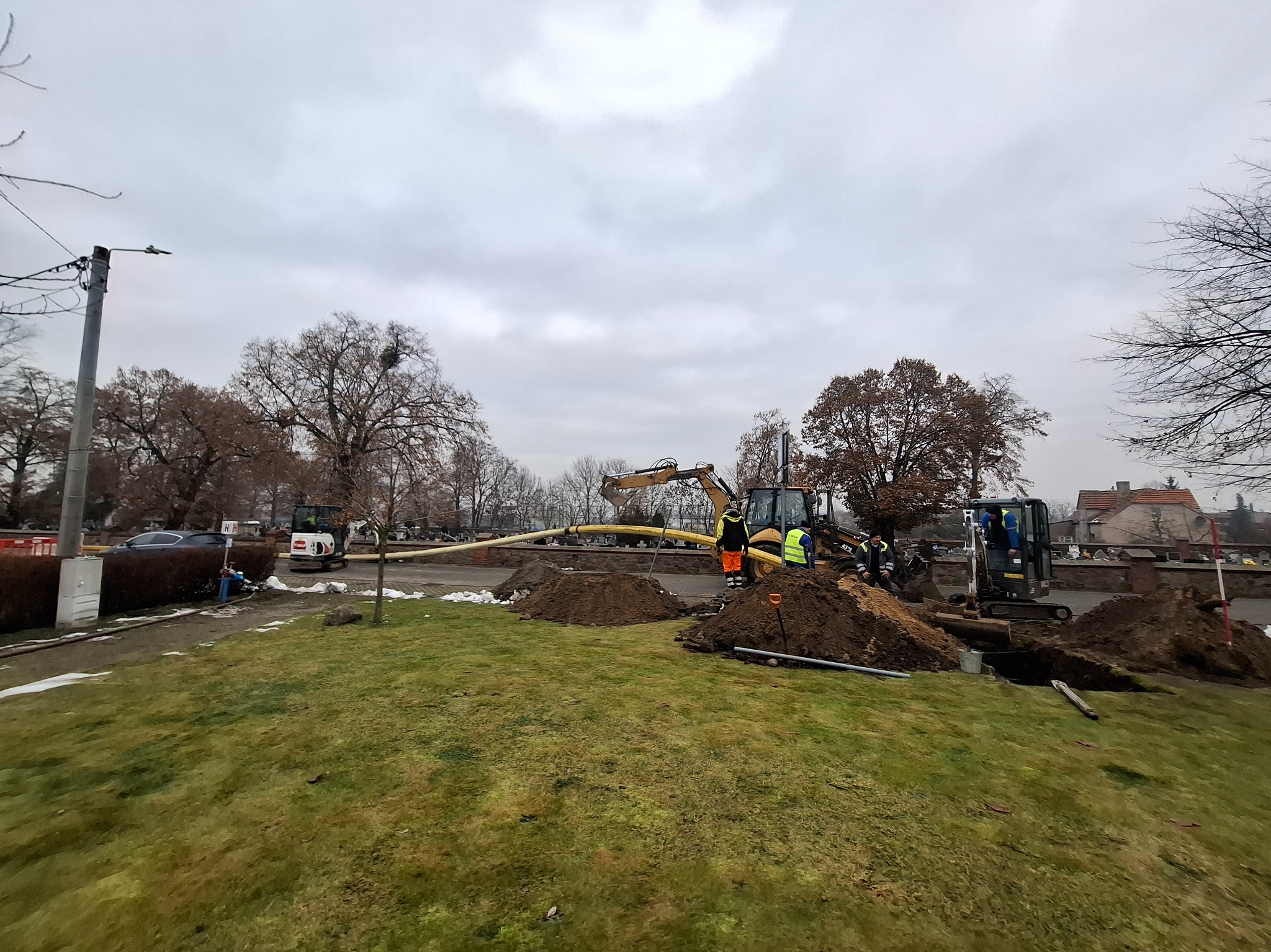 Prace ziemne z użyciem minikoparek i rur na trawniku, widoczni pracownicy w odblaskowych ubraniach. Pochmurne niebo, drzewa bez liści w tle.