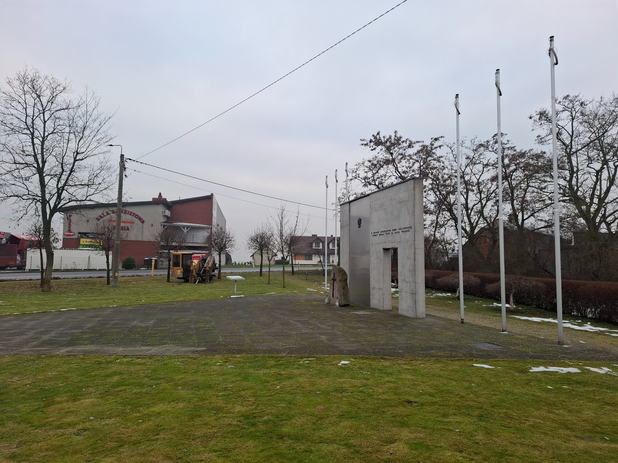 Żółta maszyna do robót ziemnych w tle pomnika z trzema masztami flagowymi i budynkiem z szyldem w tle, w pochmurny dzień.