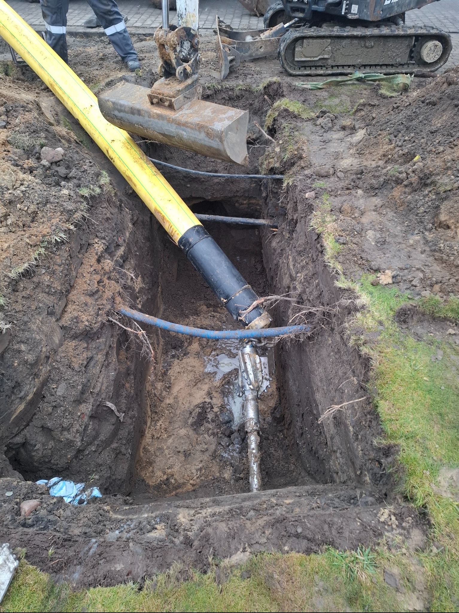 Żółta rura w wykopie z maszyną do przewiertów sterowanych. Widoczny fragment koparki i ziemia.