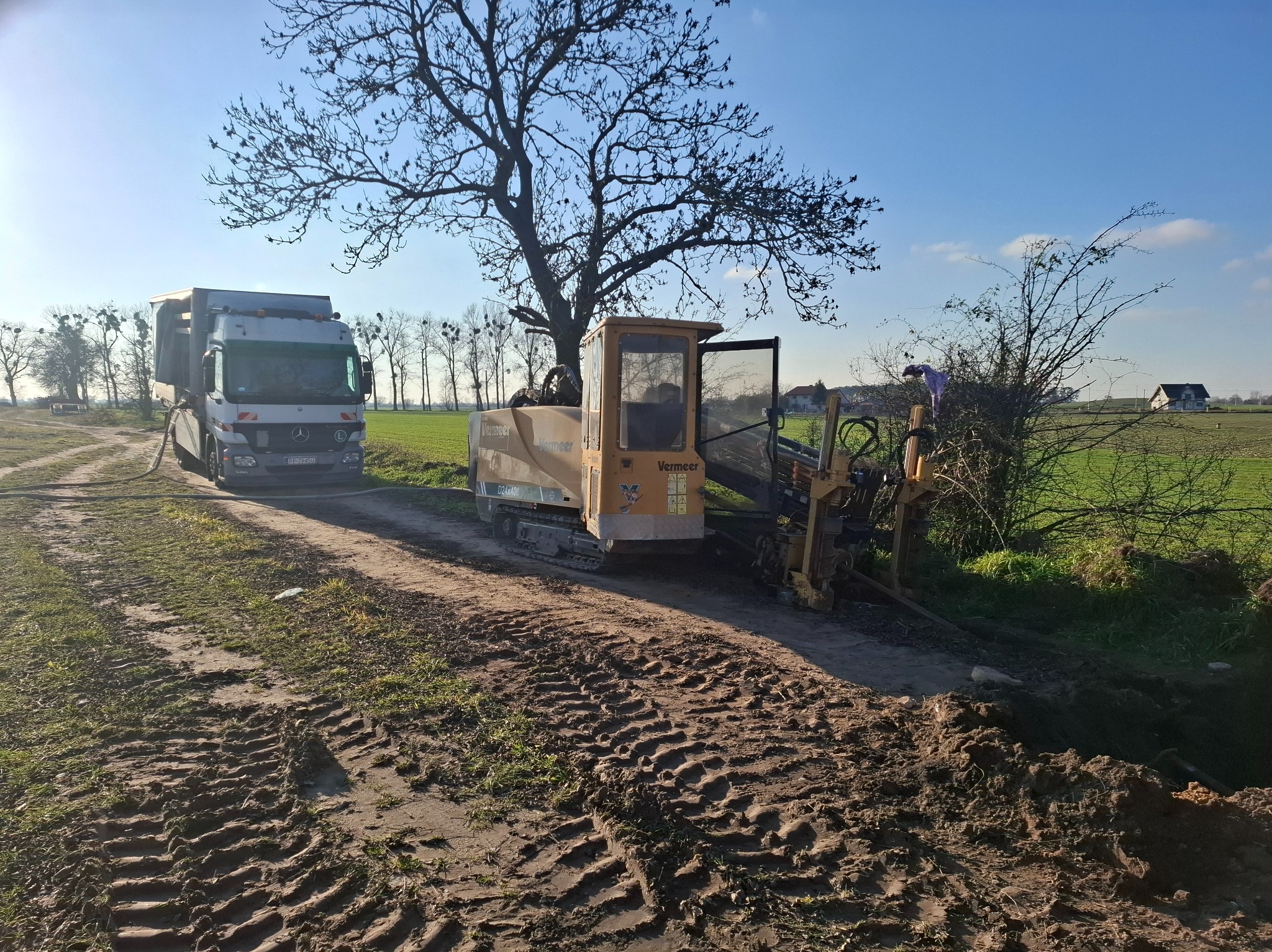 Żółta maszyna Vermeer D24x40II do robót ziemnych obok ciężarówki na tle pola i drzew w słoneczny dzień. Ślady opon na ziemi. Prace ziemne w toku.