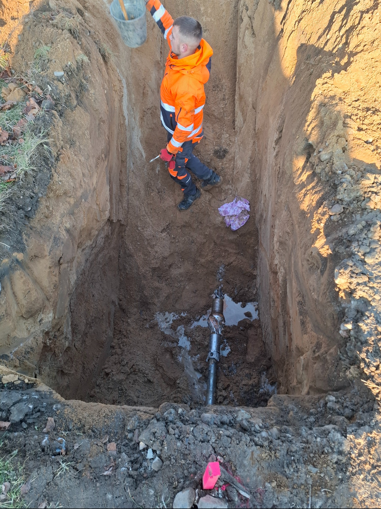 Pracownik w pomarańczowym ubraniu w głębokim wykopie z rurą. Widoczny kubeł i narzędzia. Przewiert sterowany w trakcie realizacji.